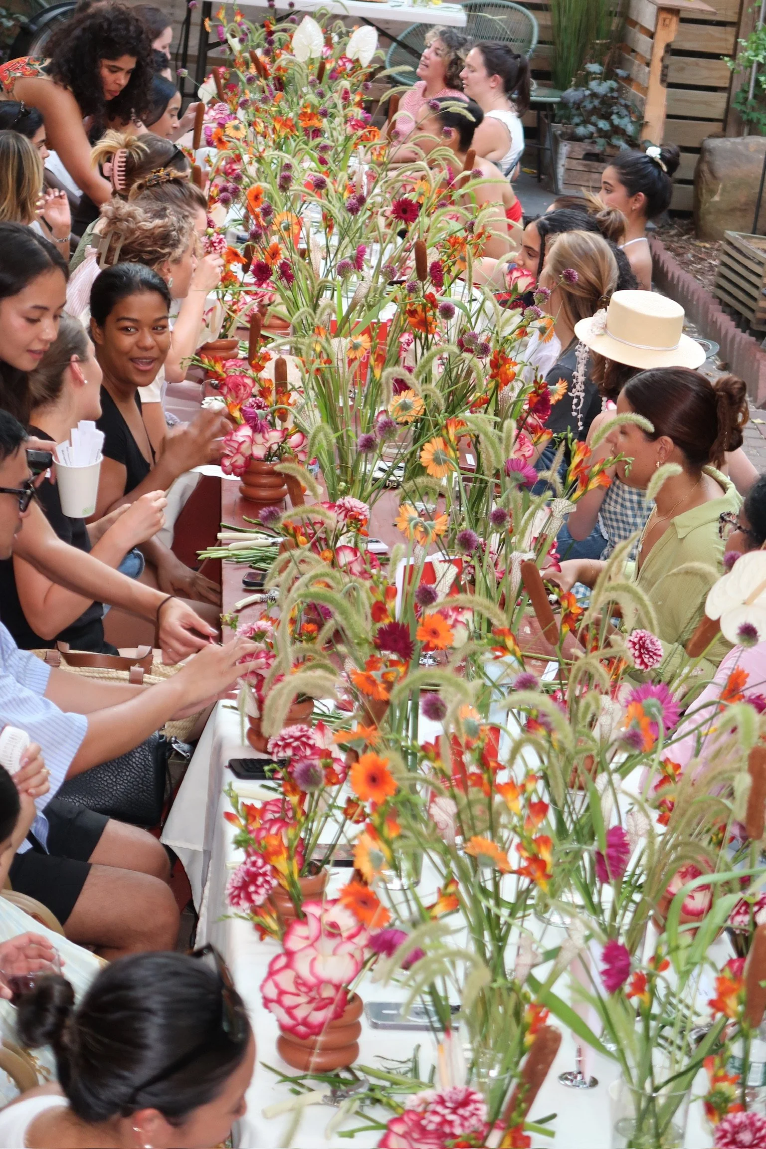 A large gathering of women sitting at a long table filled with colorful flowers in vases, engaged in conversation and smiling, in an outdoor setting with plants and wooden decor in the background.