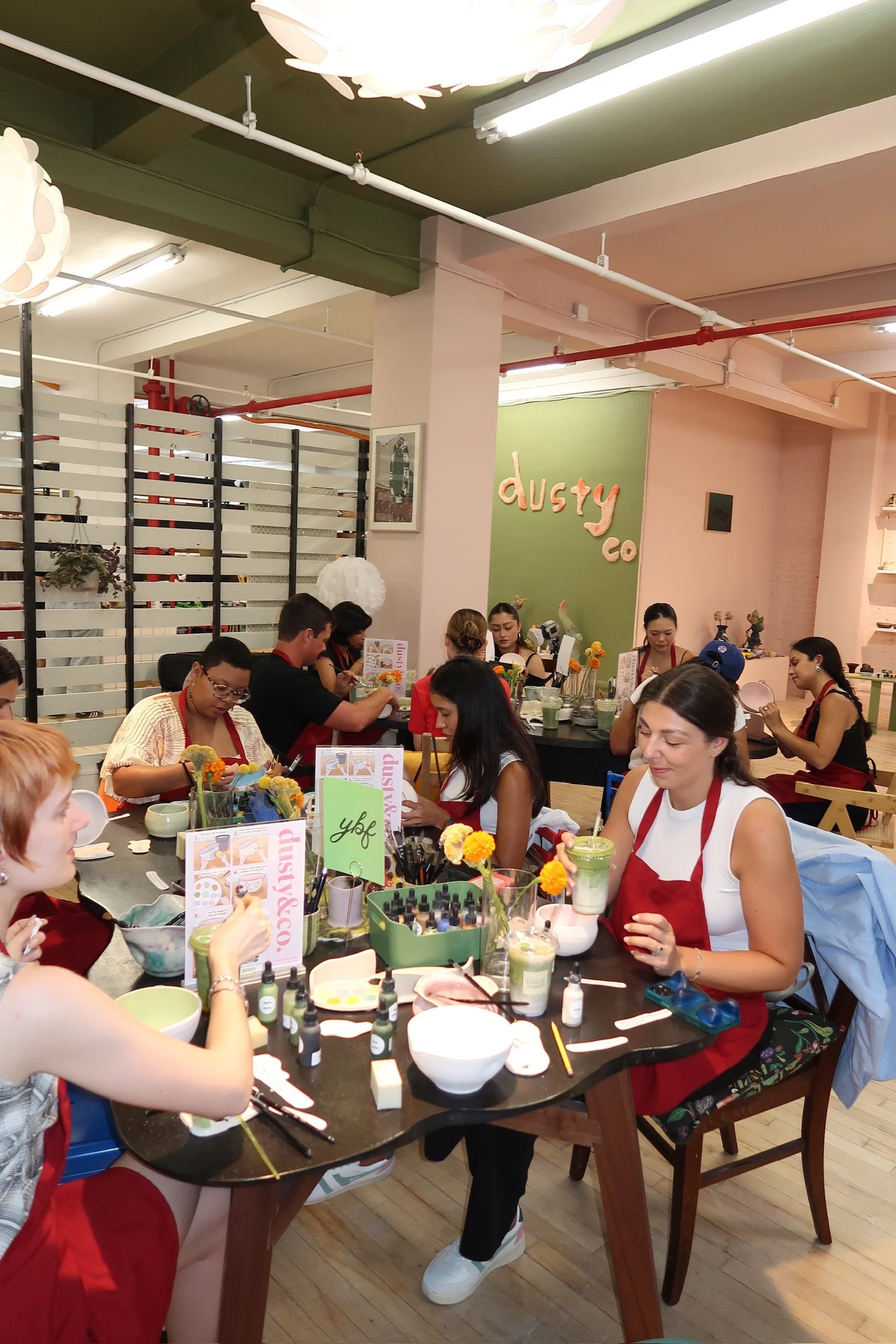 Group of women seated at a table in a nail art class, practicing nail painting and decoration, with various supplies and colorful flowers on the table, inside a decorated studio with a green wall and the sign 'dusty co' in the background.