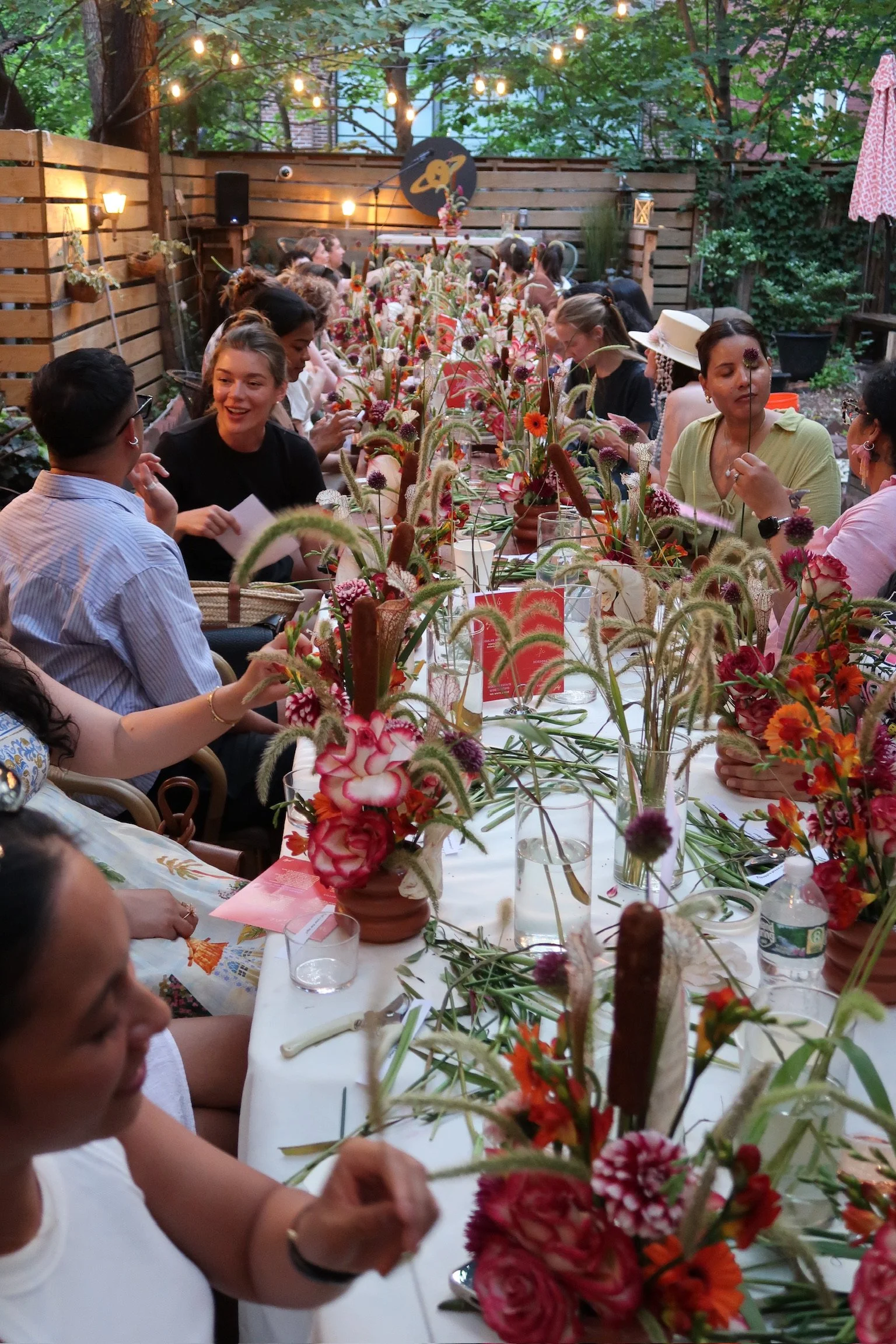 A large outdoor gathering of people seated at a long table decorated with colorful flowers, with string lights overhead and trees in the background.