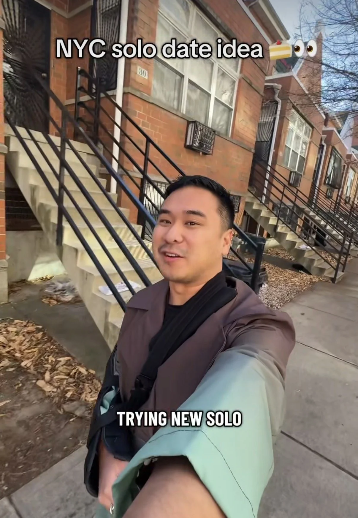 A young man taking a selfie outside a brick apartment building with staircases, with text overlay saying 'NYC solo date idea' with cake and eyes emojis, and 'TRYING NEW SOLO'.