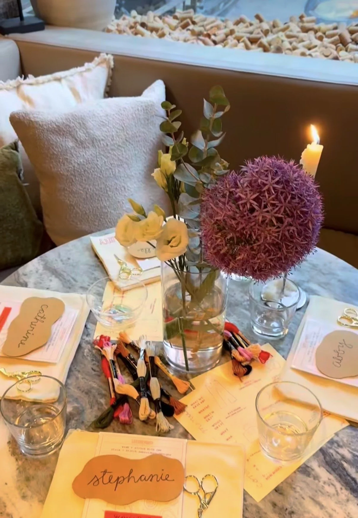 Decorative table setting with a floral centerpiece featuring a pink allium, white roses, and eucalyptus leaves in a glass vase, surrounded by glasses, scissors, and name tags for an event or gathering.