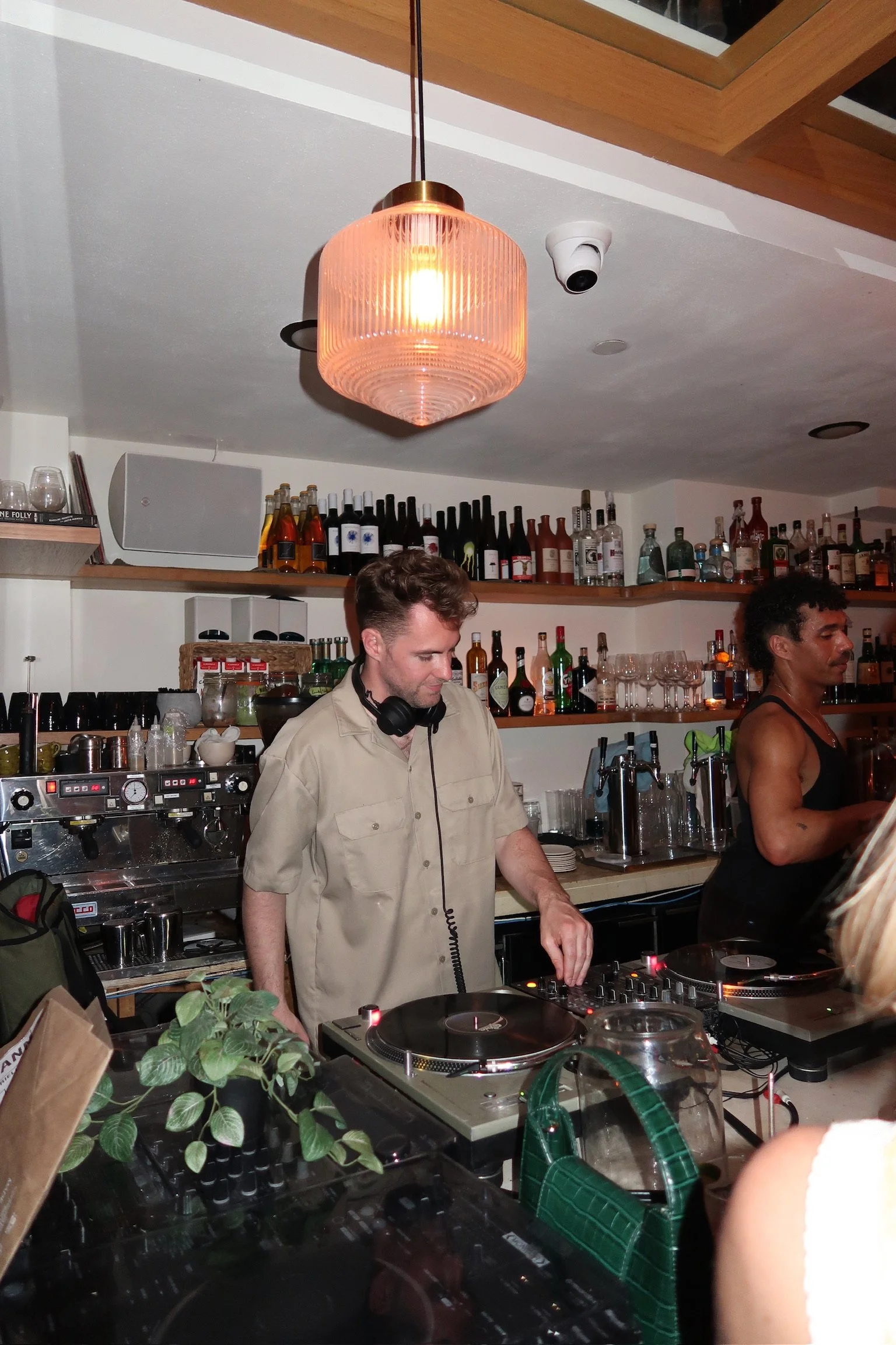 A DJ with headphones and a beige shirt is playing music in a bar or lounge. The scene includes bottles of alcohol, glassware, and a chandelier overhead.