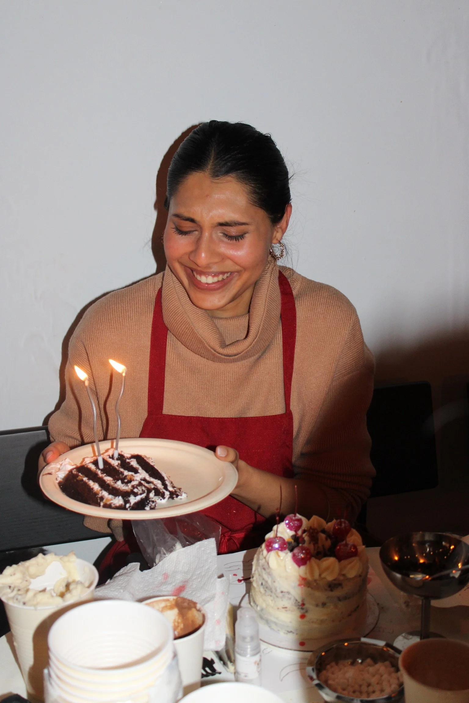 A woman with dark hair smiling with closed eyes, holding a plate with a chocolate cake slice and lit candles, at a celebration with various desserts on a table.