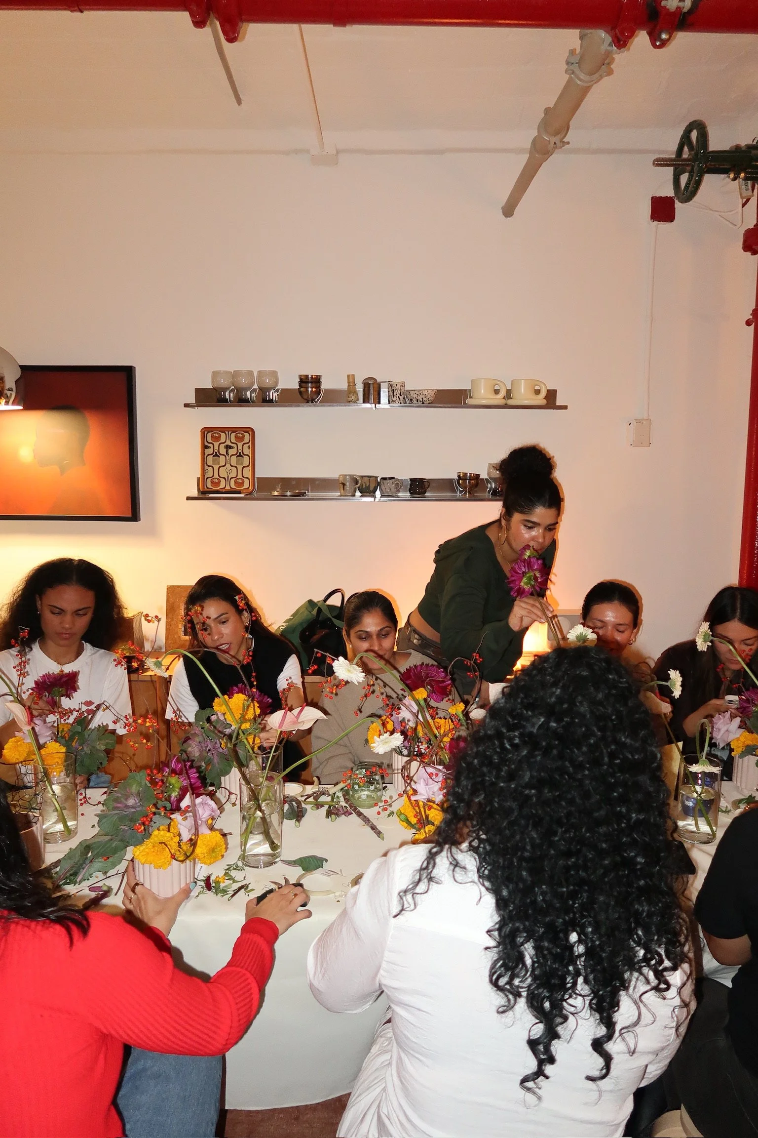 Group of women seated at a table with floral arrangements, while one woman arranges flowers in a vase; the setting appears to be an indoor workshop or gathering.