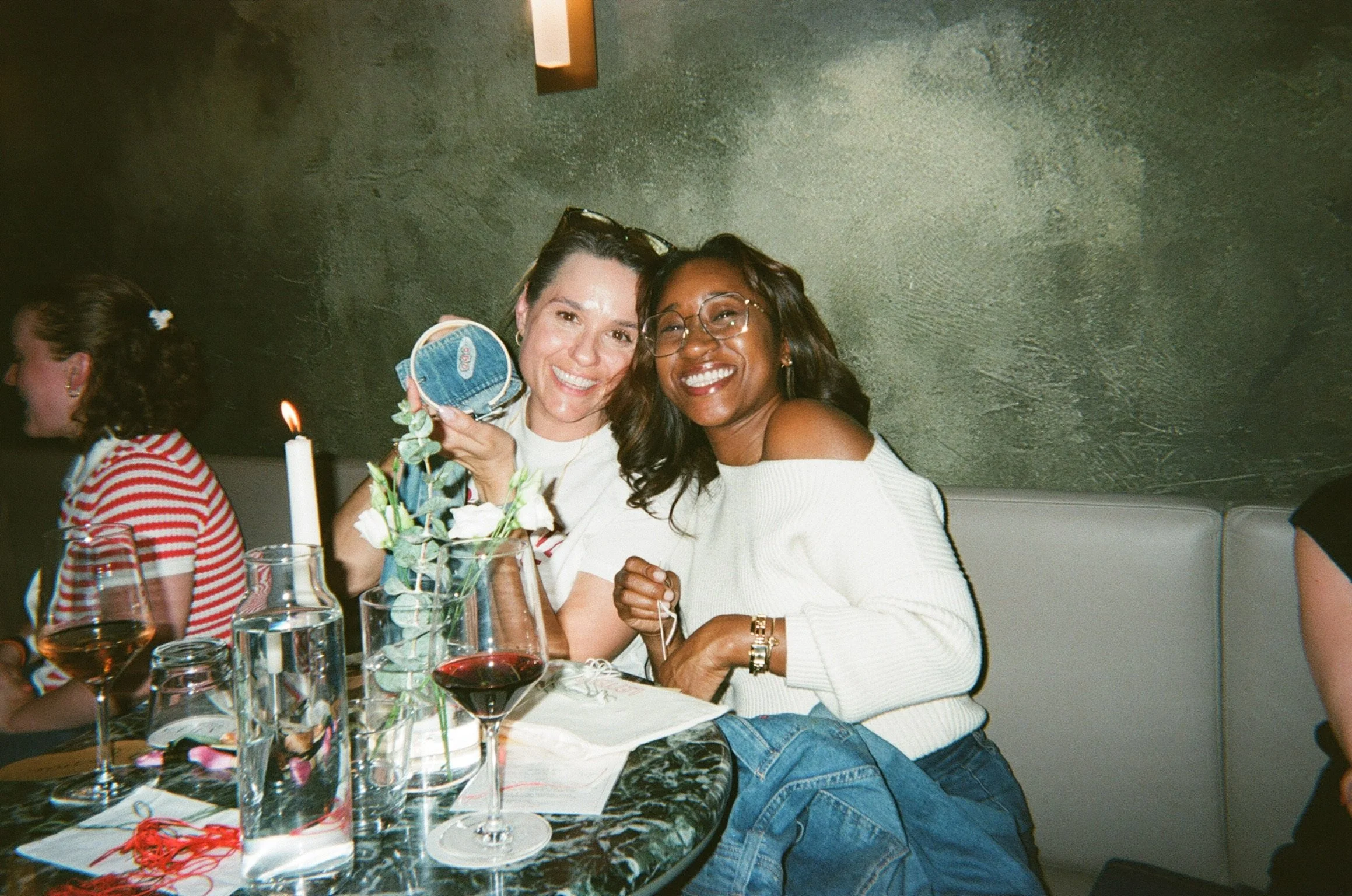 Two women smiling at a dinner table with drinks and candles, celebrating with gifts in a dimly lit restaurant.