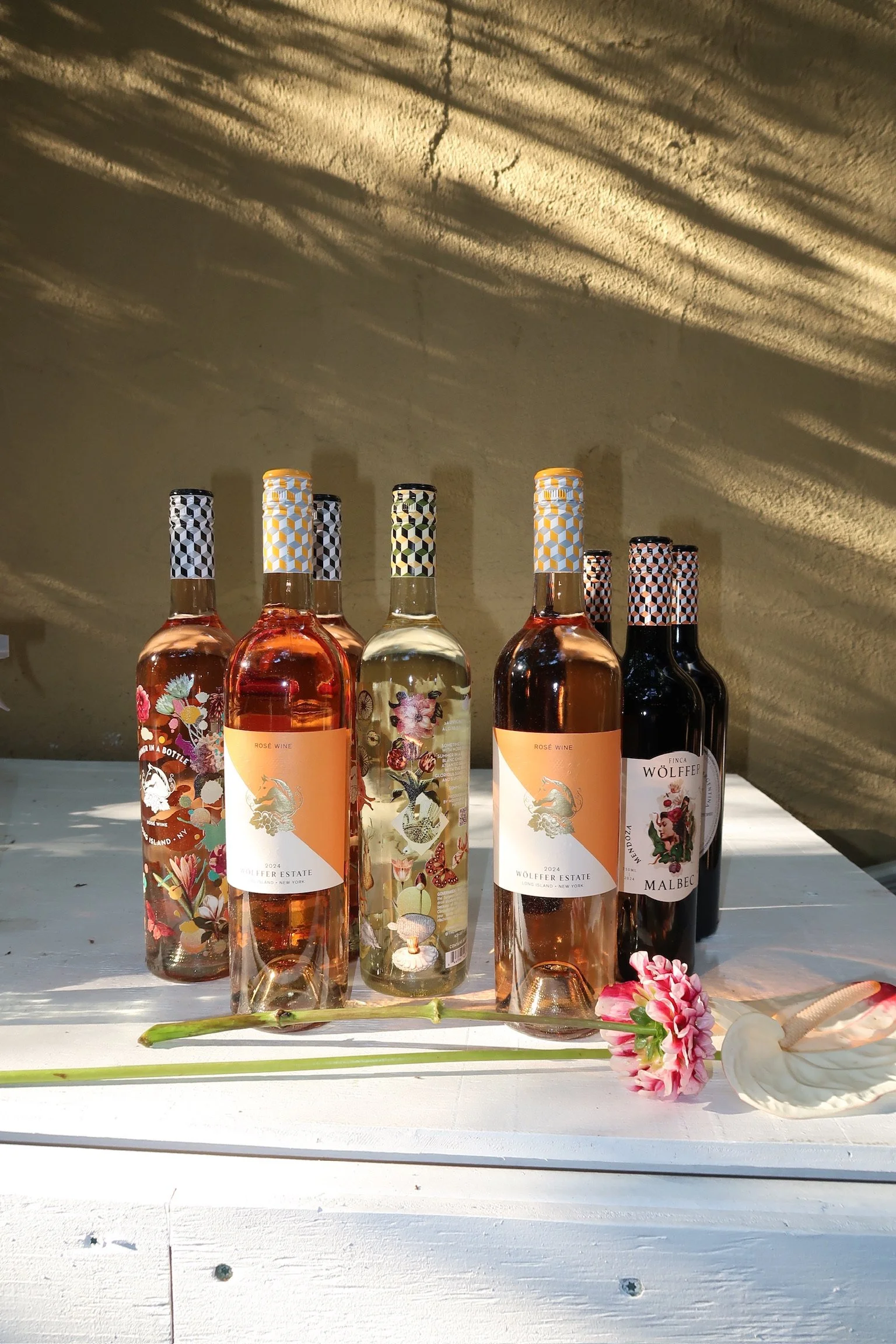 Six bottles of wine, five pink and one black, are arranged on a white table with two flowers in front of them, against a yellow wall with leopard print shadows.