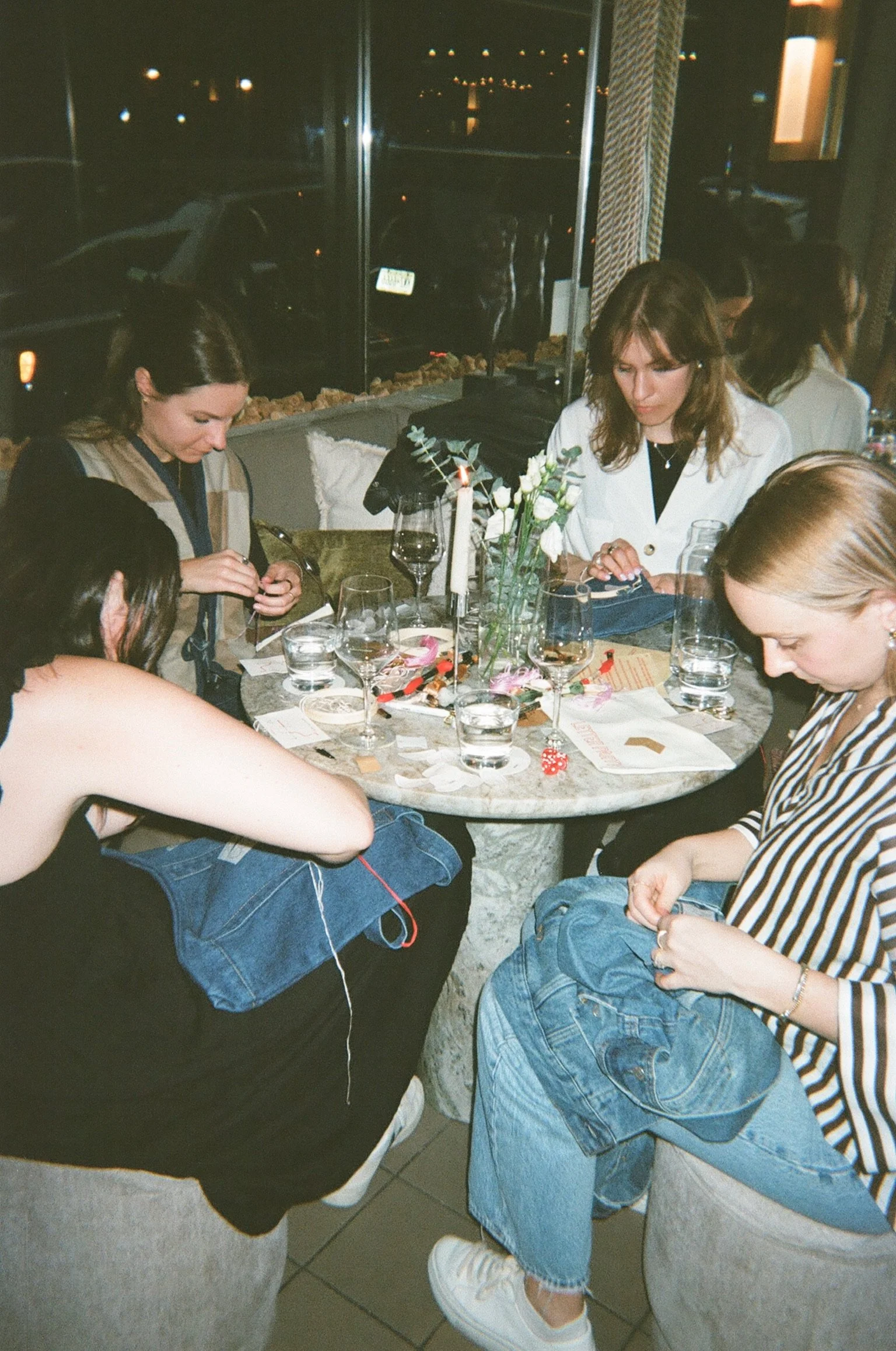 A group of young women sitting around a table in a dimly lit restaurant, engaged in knitting or crafting, with glasses and a floral centerpiece on the table.