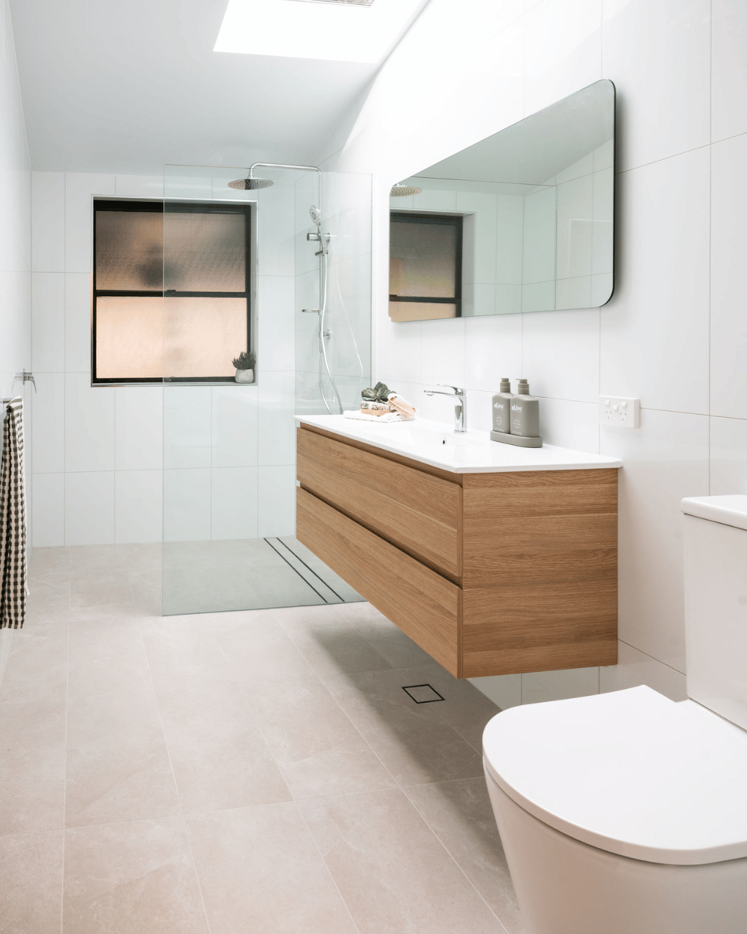 Modern bathroom with white tiled walls, a floating wooden vanity with a white countertop, and a large mirror. There is a walk-in shower with a glass partition, a window with frosted glass, and a toilet.