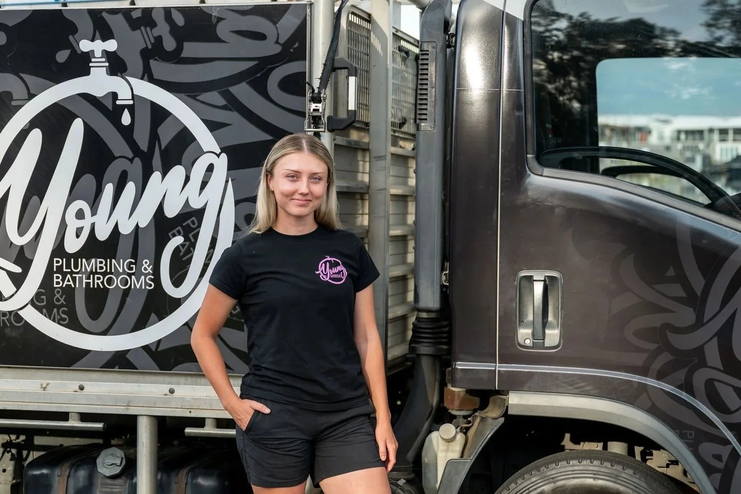 A woman in a black T-shirt standing next to a truck with the words "Young Plumbing & Bathrooms" on the side, with her hand in her pocket.