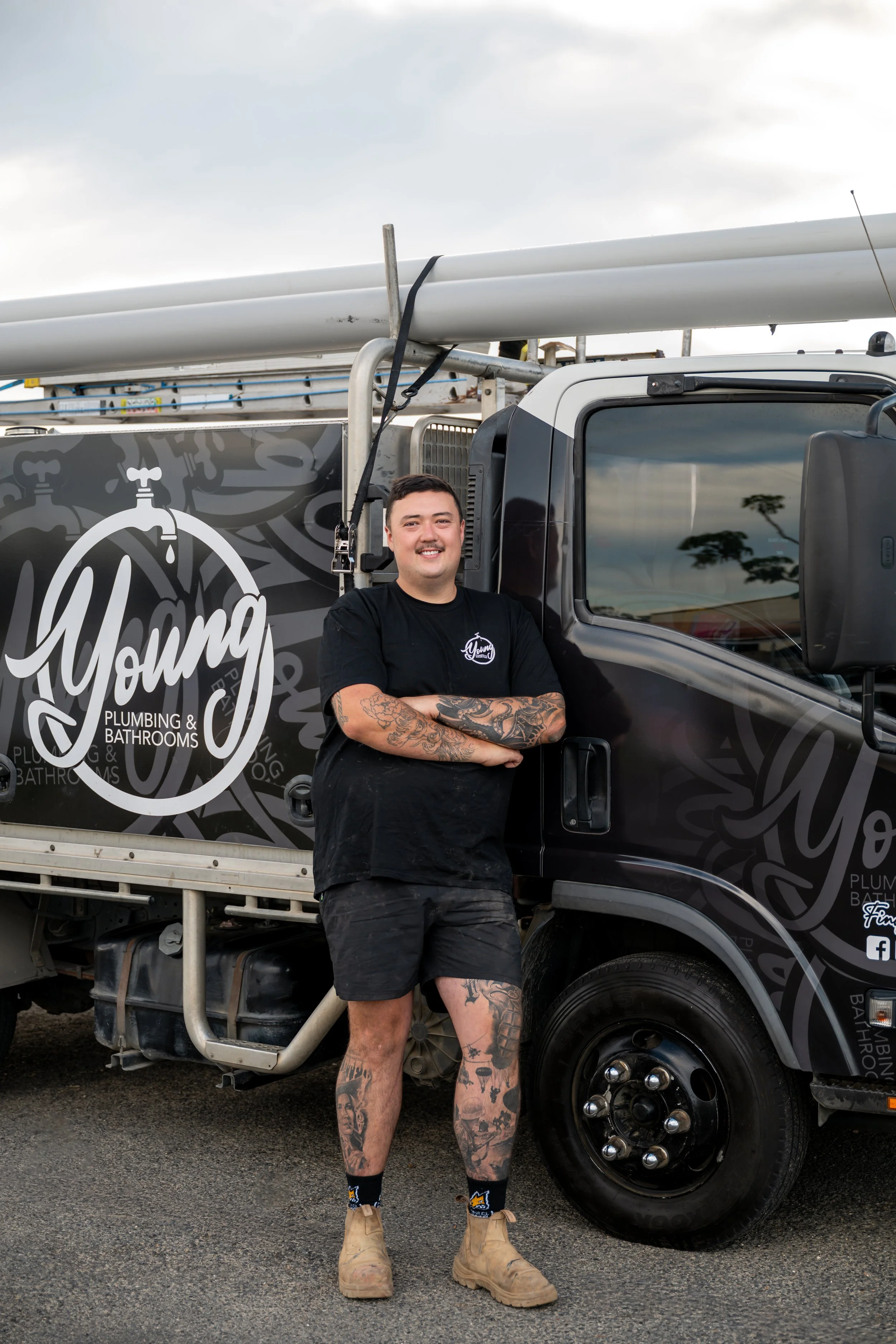 A man with tattoos on his arms and legs, wearing a black t-shirt, shorts, and tan work boots, stands next to a plumbing truck with a logo that reads 'Young Plumbing & Bathrooms.' The man is smiling and crossing his arms, with blue sky and some trees reflected in the truck's window.