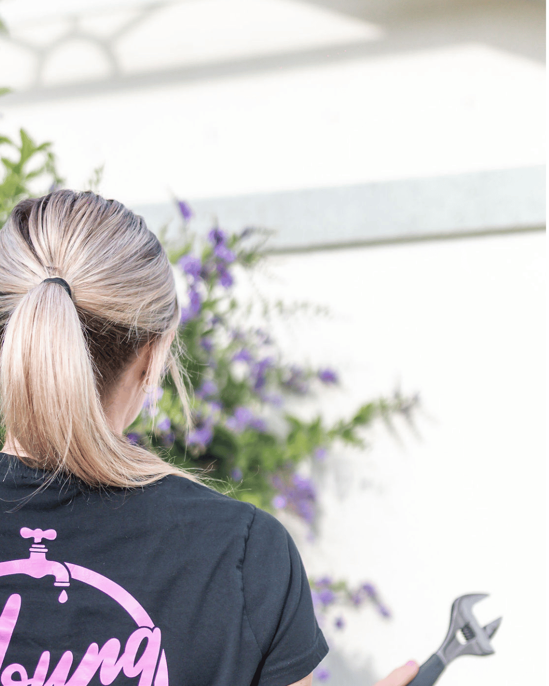 A woman with blond hair tied in a ponytail holds a wrench, facing away, with purple flowers and a white wall in the background.