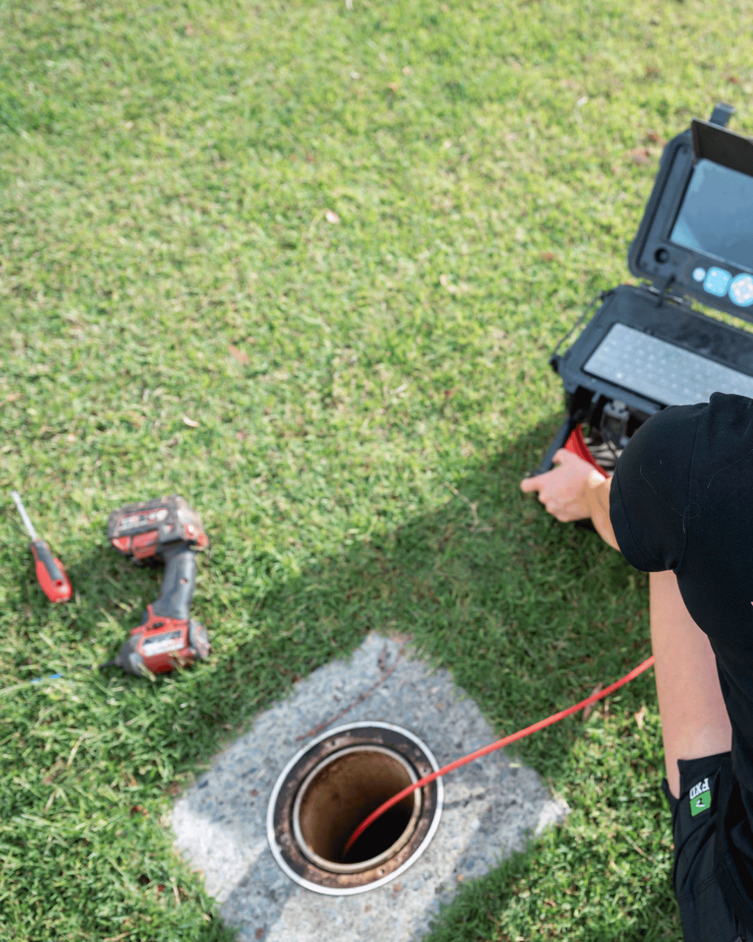 A person inspecting a sewer manhole using testing equipment on a grassy area. Tools like a screwdriver and a cordless drill are nearby.