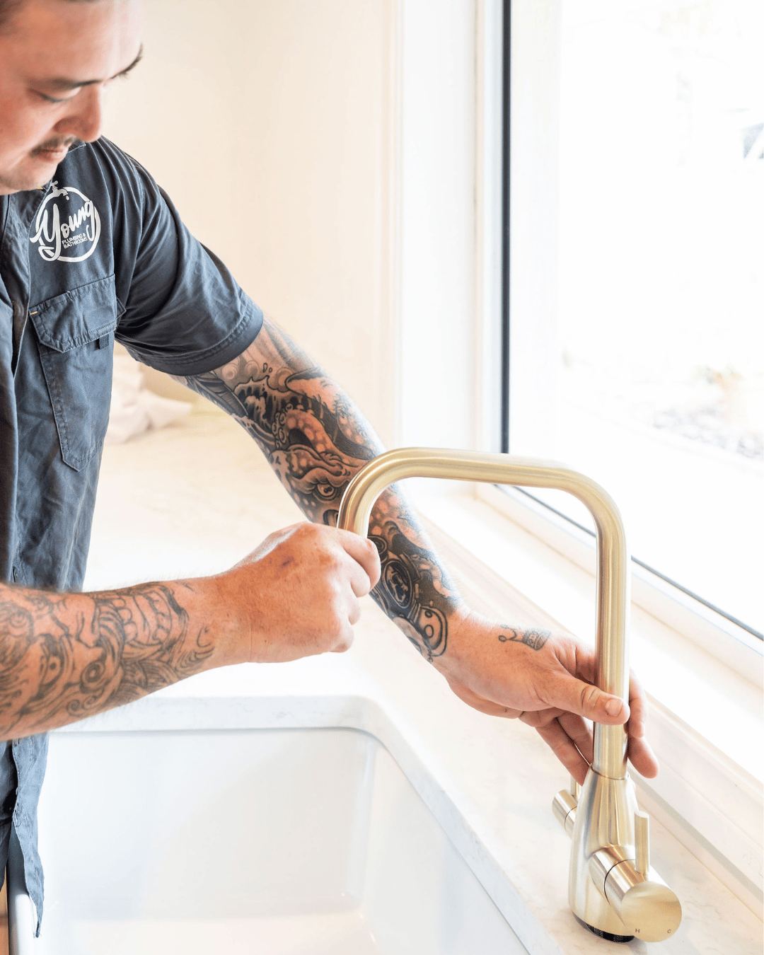 A man with tattoos on his arms turning on a gold-colored kitchen faucet in a bright kitchen near a window.