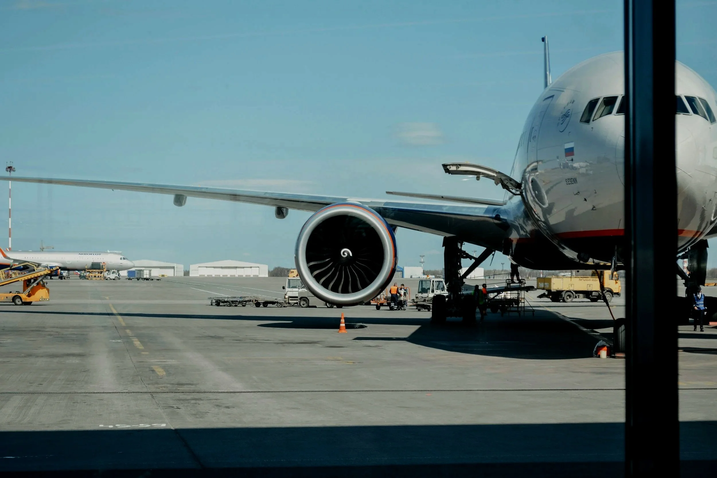 Front view of a commercial airplane parked at an airport gate, with ground crew and luggage vehicles nearby, seen through a window.