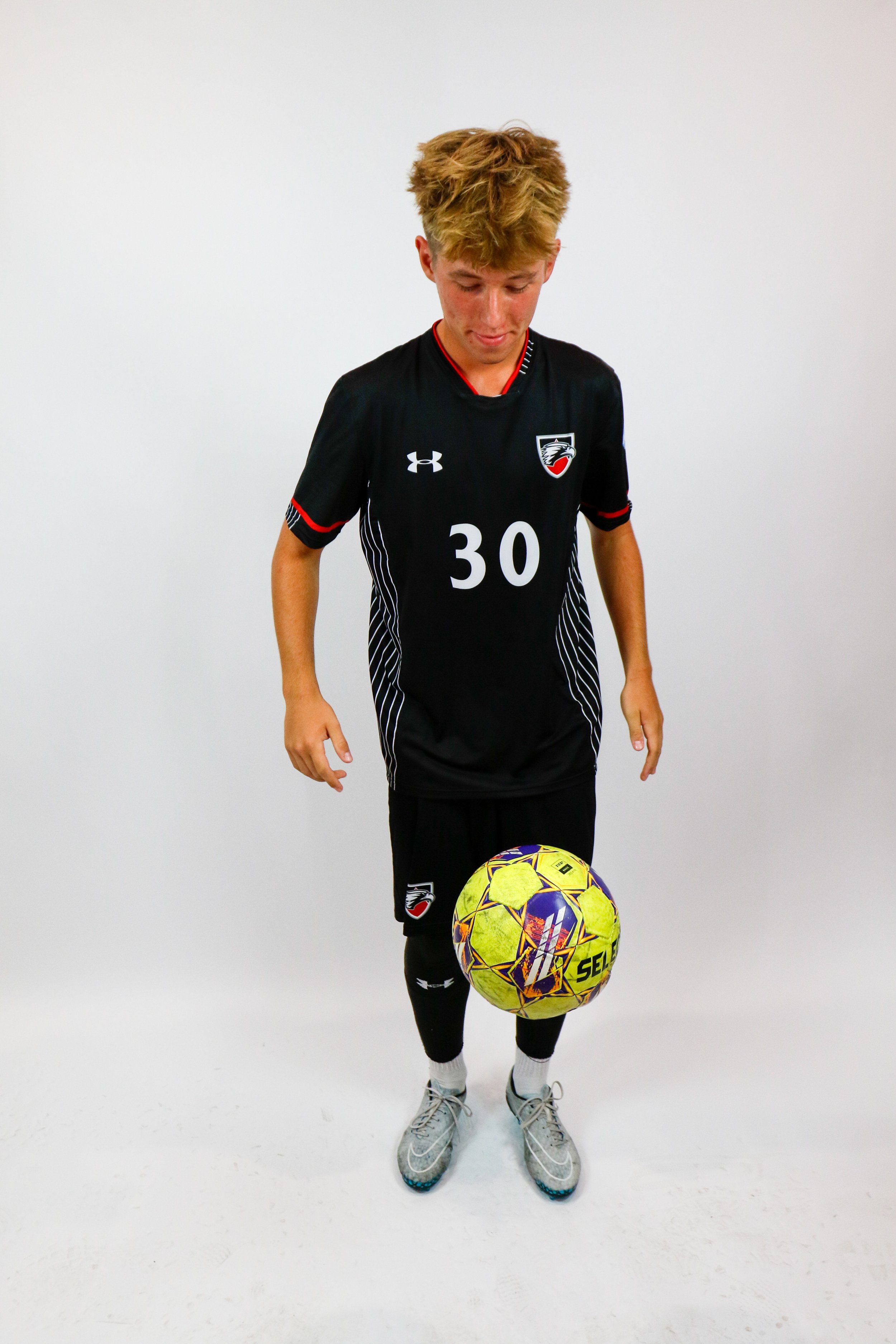 A young male soccer player wearing a black uniform with the number 30, standing on an all-white background, balancing a yellow and purple soccer ball on his right foot.