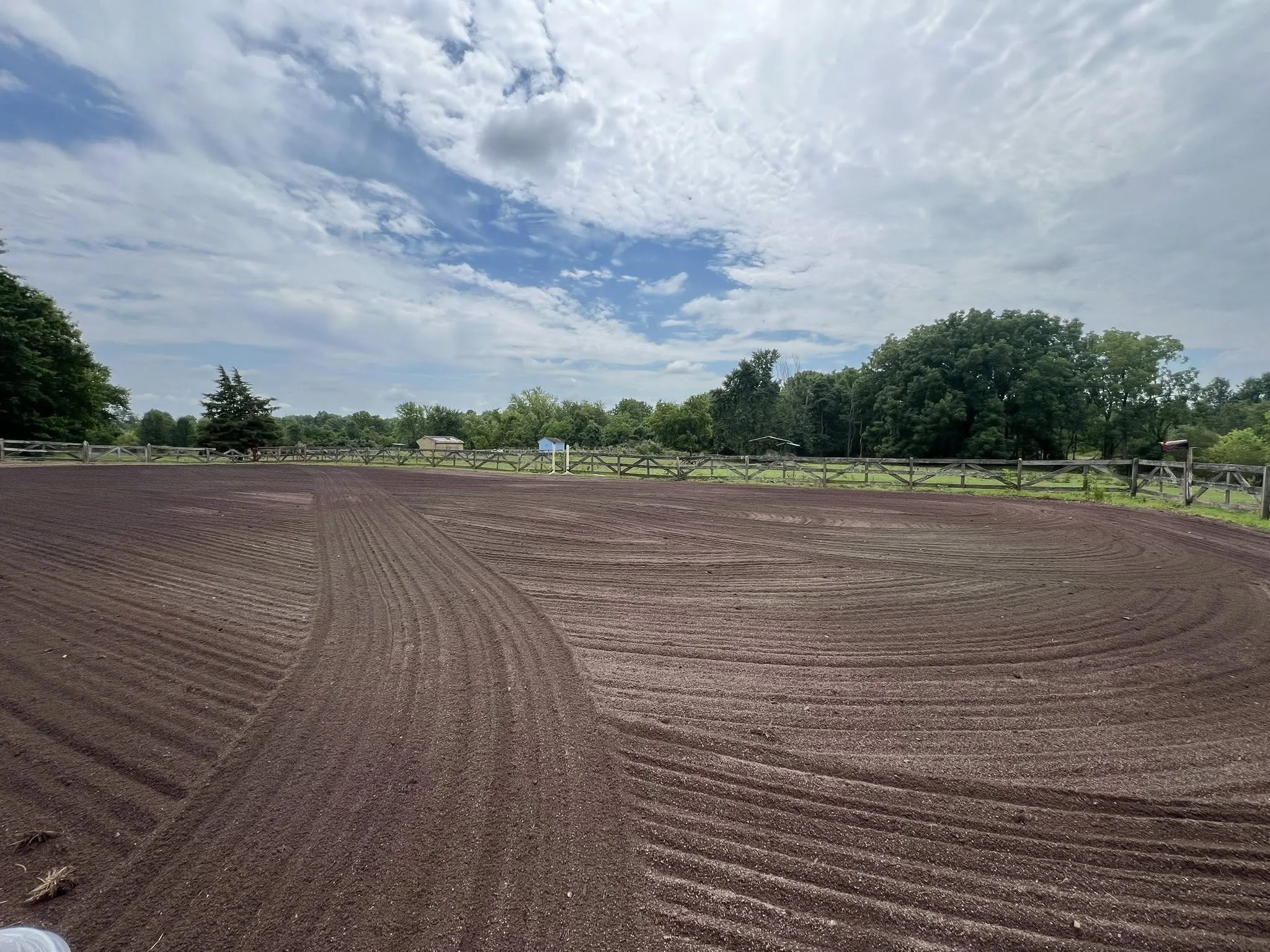 An empty, freshly tilled horse riding arena with visible grooming lines, surrounded by a wooden fence, under a partly cloudy sky with patches of blue.