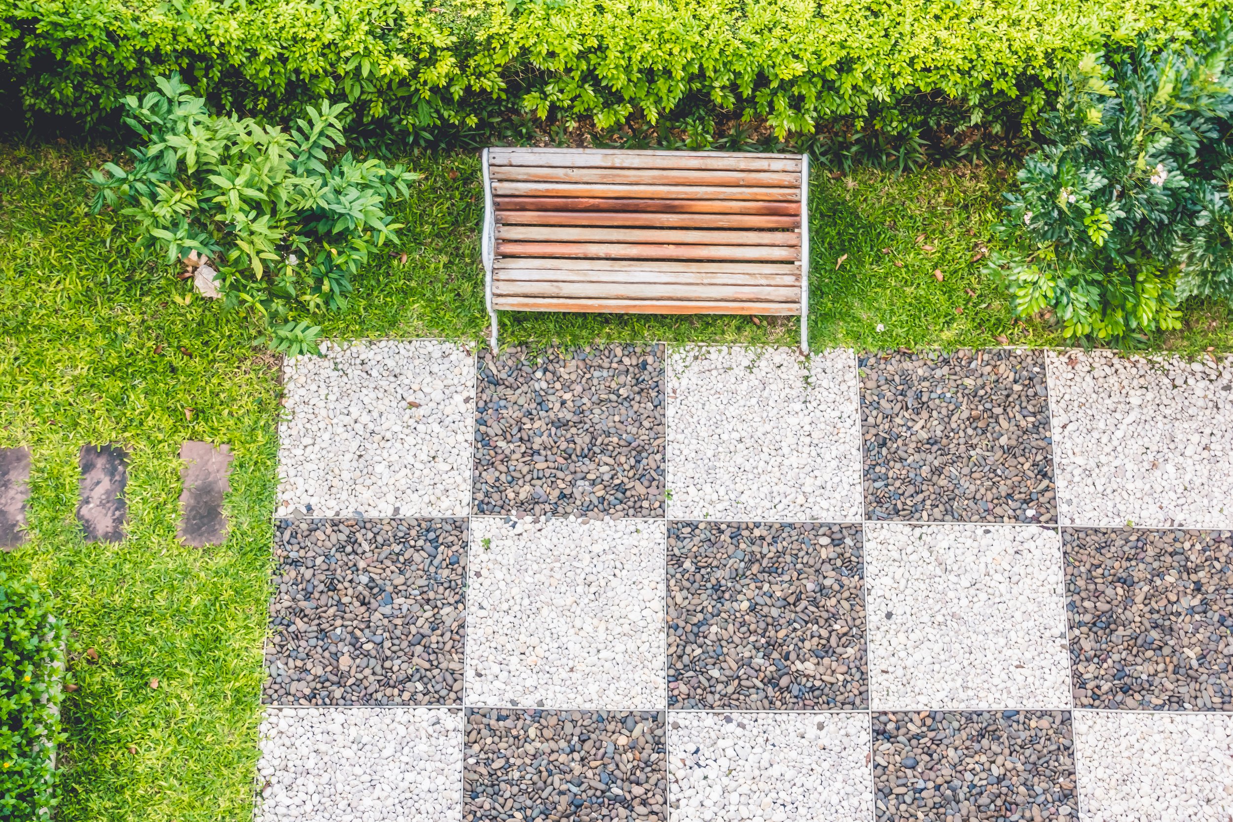 Top-down view of a garden with a wooden bench and a patchwork of tiled and pebble paths surrounded by various green bushes and plants.