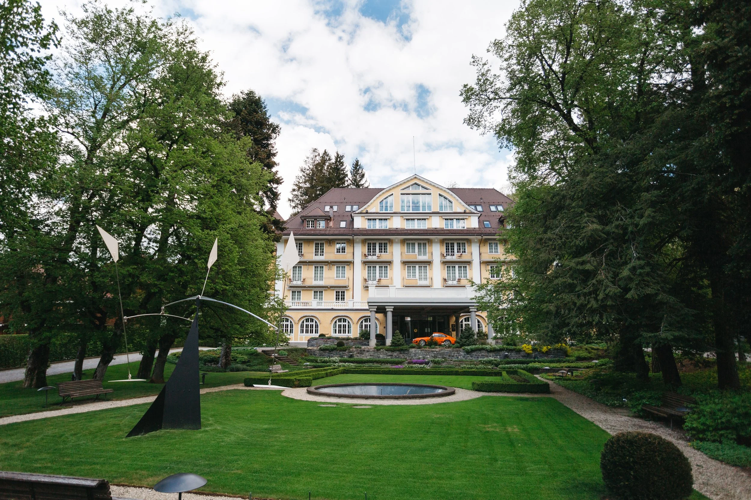 View of a large yellow building resembling a hotel or mansion, surrounded by a well-maintained garden with trees, grass, a fountain, and sculptures, under a partly cloudy sky.