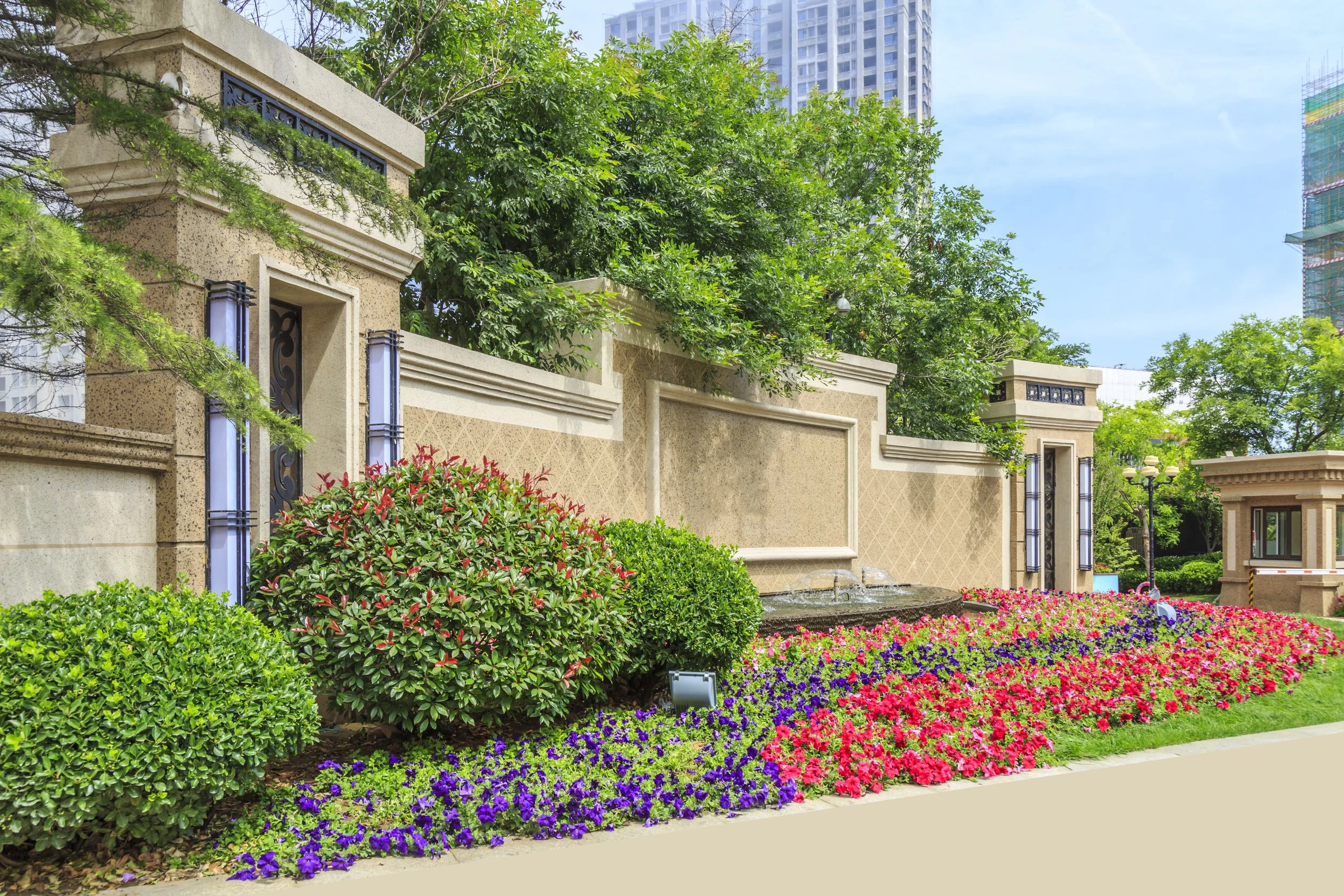 A landscaped area with colorful flowers including pink, purple, and red, green bushes, and trees. There is a decorative beige stone wall with black metal accents and modern light fixtures. Behind the greenery are tall buildings and a partly cloudy sk