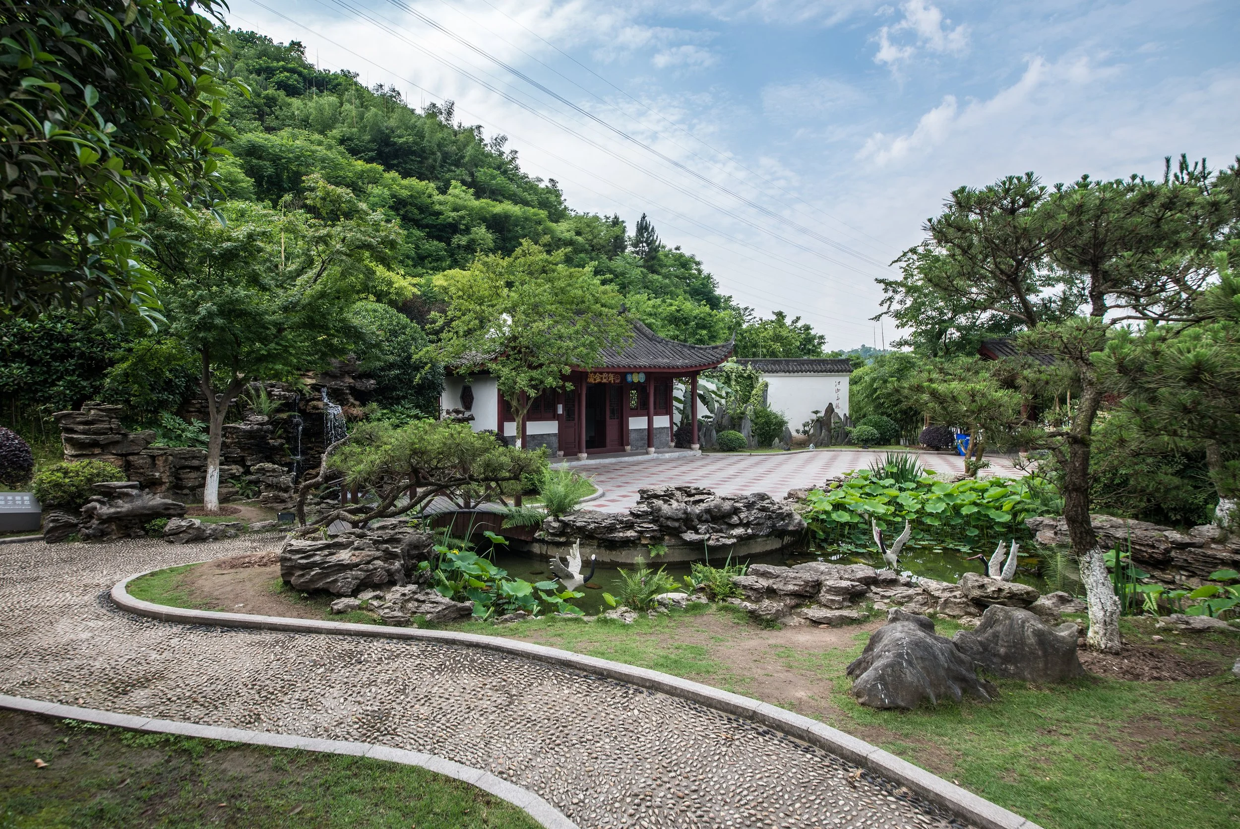 Traditional Chinese garden with a pond, stone rocks, lush green trees, and a small pavilion with curved rooftop, set against a hillside with dense forest and power lines in the background.