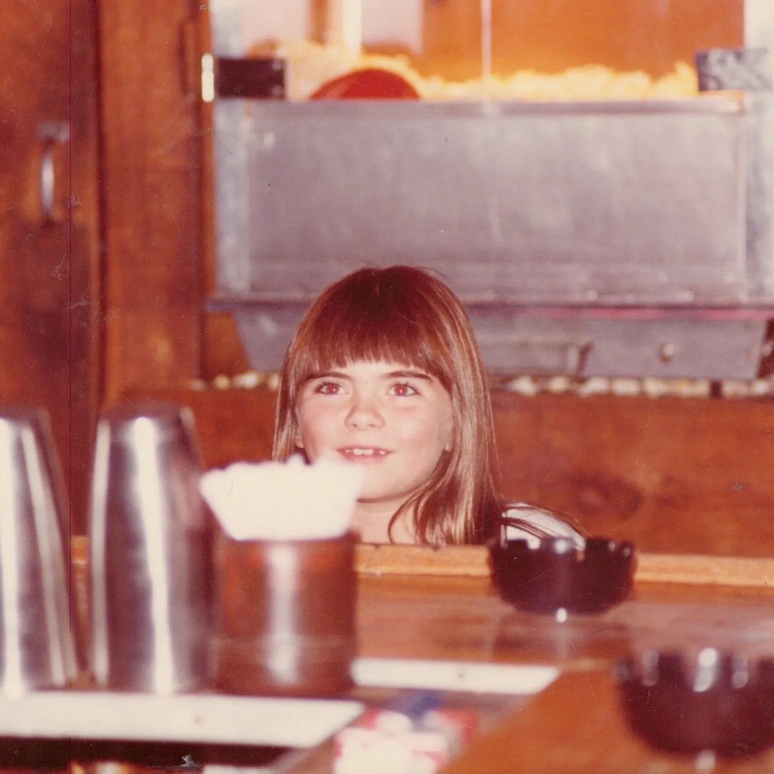 A young girl with reddish-brown hair and a big smile sitting at a table in a diner or restaurant, with food items and utensils on the table.