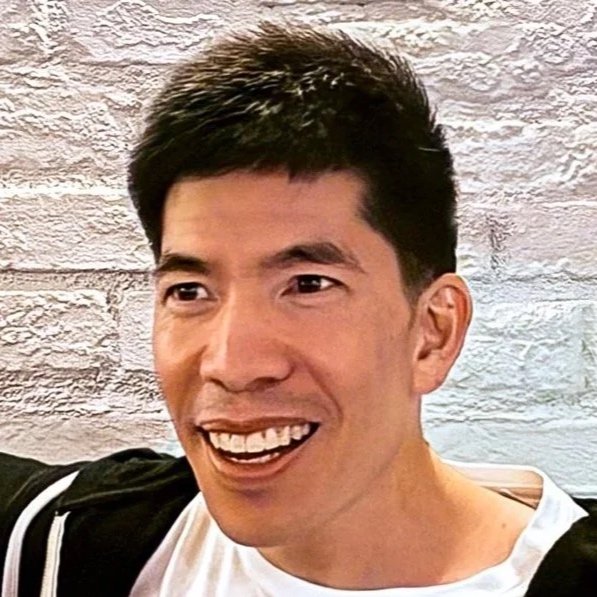 Close-up of a smiling man with short black hair, wearing a white shirt and black jacket, in front of a white brick wall.