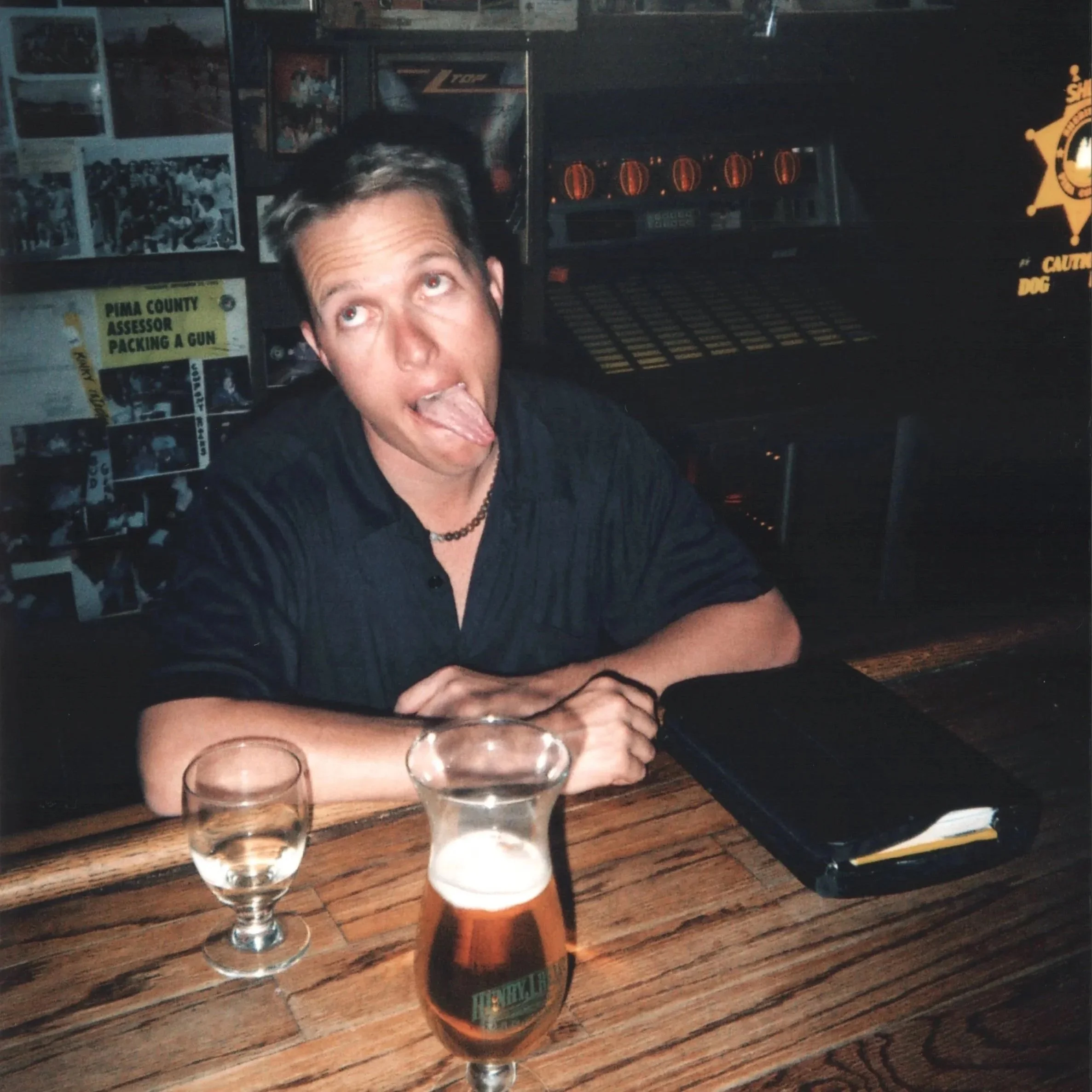 A person with short hair and a necklace sticking out their tongue at a bar or pub with four shot glasses and beers on a wooden table.