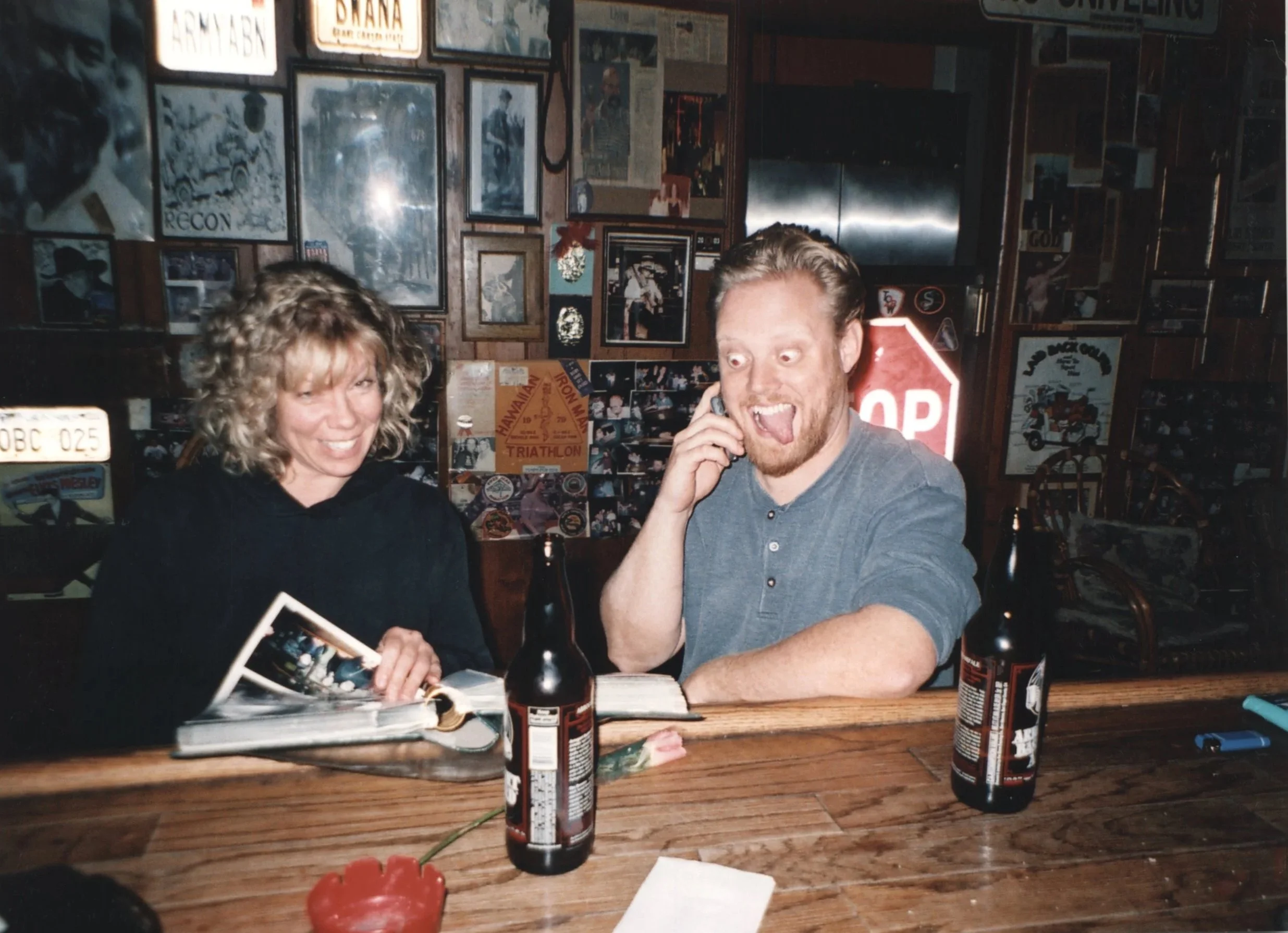 A woman with curly blonde hair and a man with red hair and a beard sitting at a wooden table in a dimly lit restaurant decorated with numerous framed posters and photographs on the wall. The woman is smiling, and the man is talking on a cellphone with his tongue out.