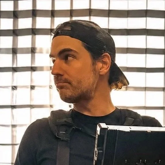 A man with long dark hair under a backward baseball cap, wearing a black shirt and backpack, looking to the side in front of a patterned window.