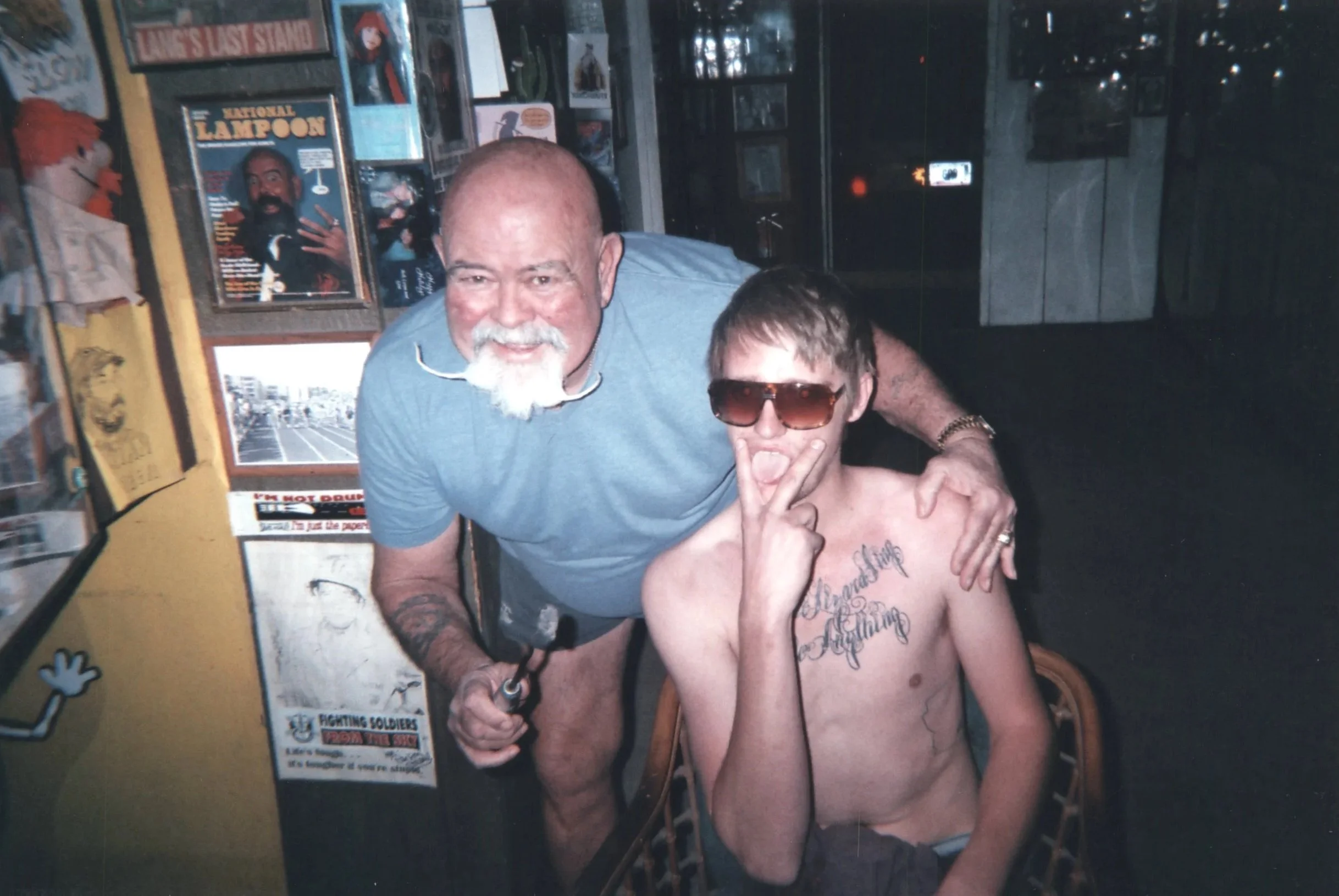 A man with a bald head and gray beard standing next to a shirtless young man wearing sunglasses and tattoos on his chest. The man with the beard has his arm around the young man's shoulders, and they are smiling and posing for the camera in a room decorated with posters and memorabilia.