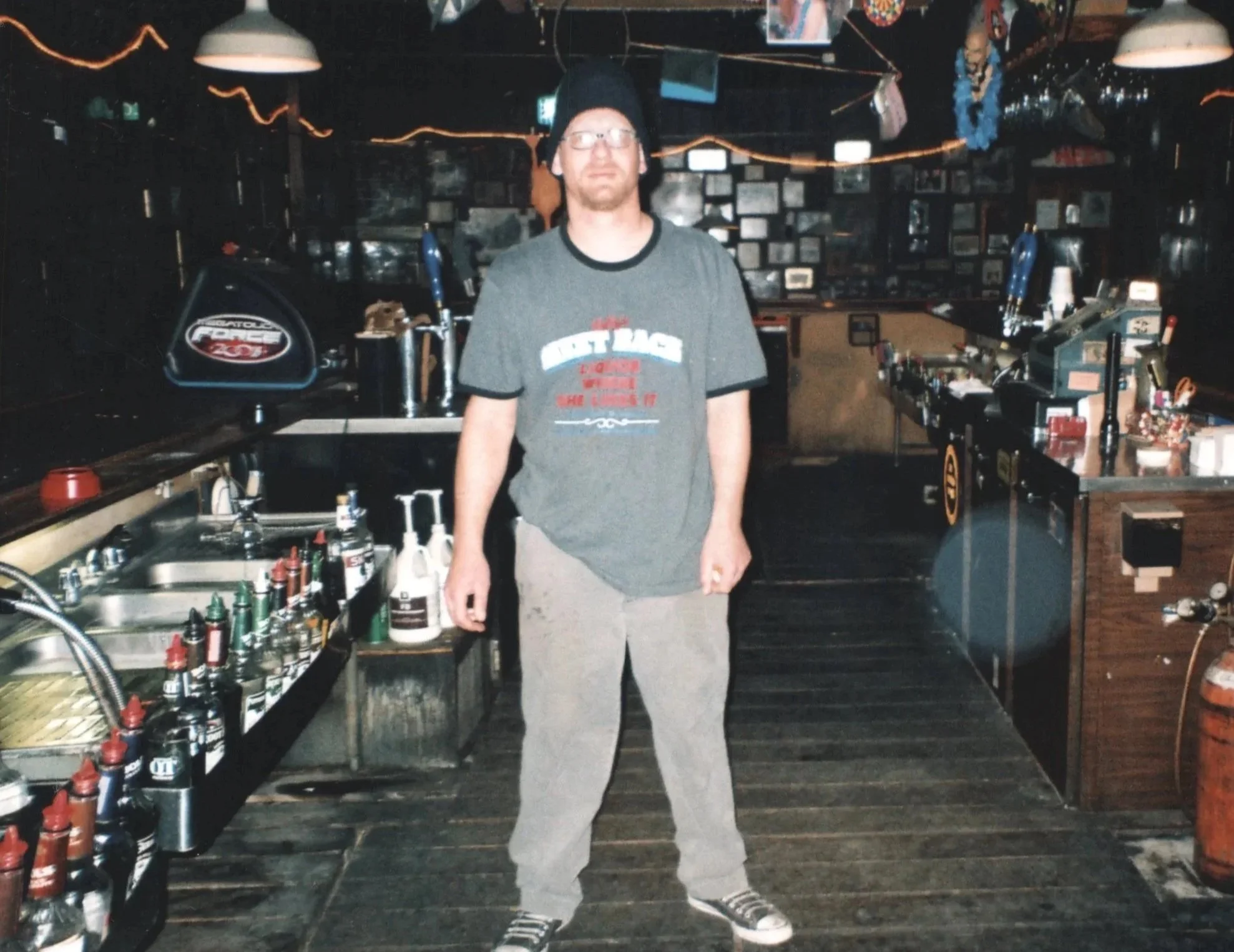 A man with glasses and a beard stands inside a bar or pub, wearing a dark beanie, gray T-shirt, and gray pants. The background is decorated with framed pictures on dark-colored walls, and there are neon lights. The bar counter behind him has bottles, glasses, and bar equipment.