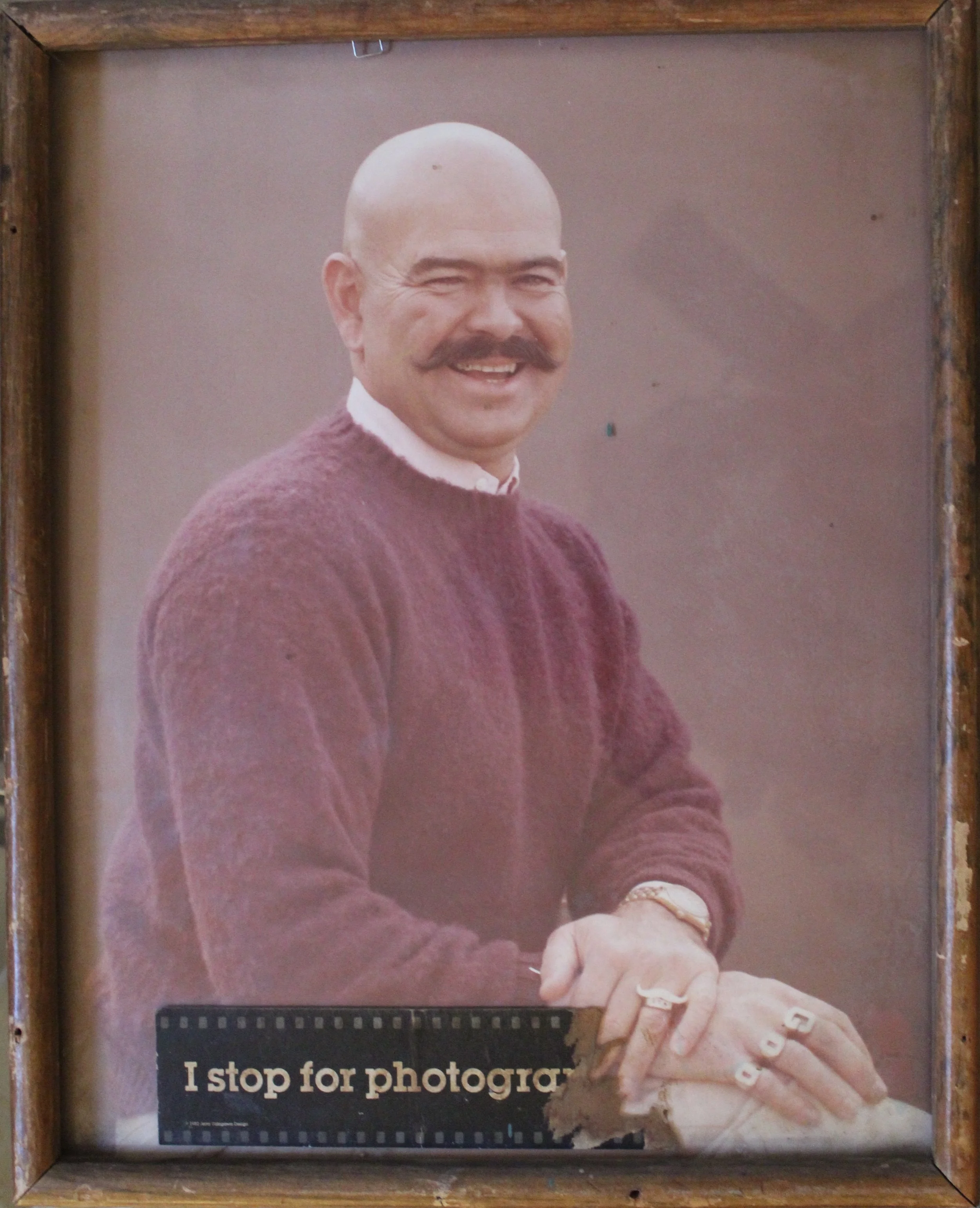 Close-up of a person with a bald head and mustache, smiling and holding a pair of white shoes. Part of a poem or text is visible at the bottom of the image.