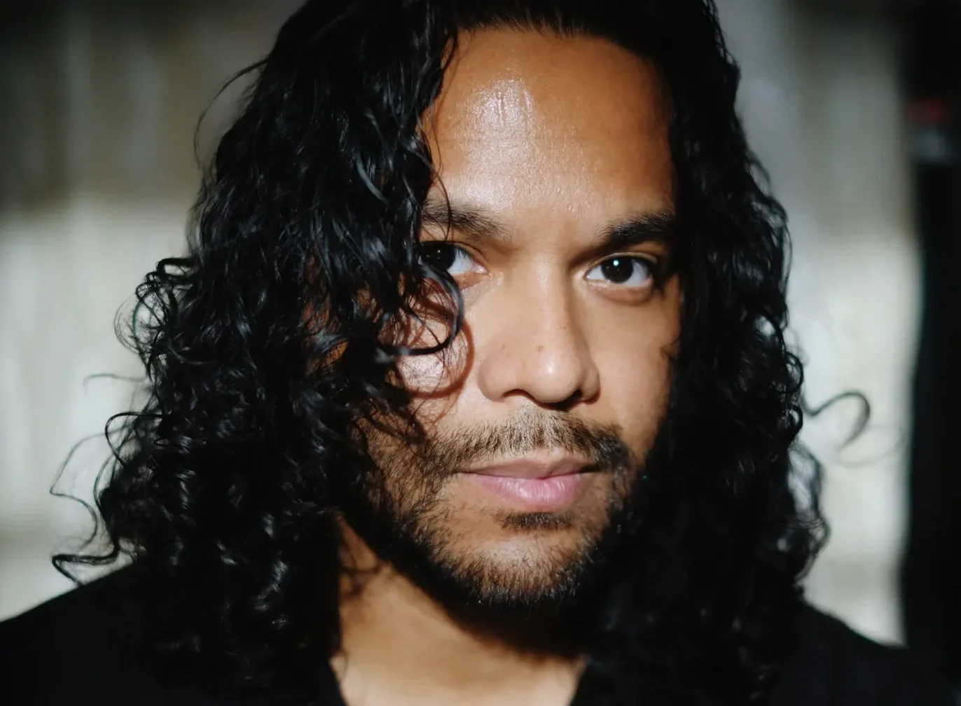 Close-up of a man with long, curly black hair and a beard, looking at the camera.