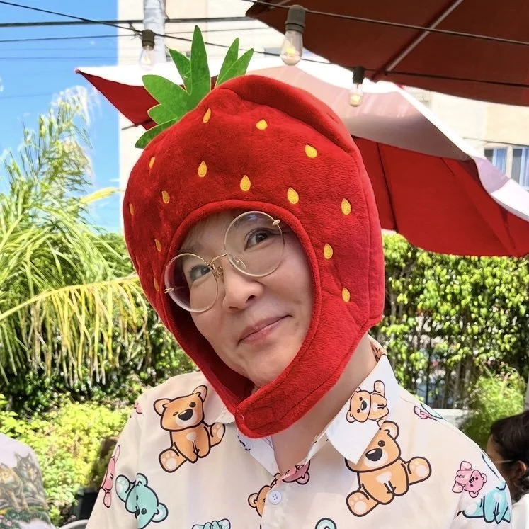 Person wearing a red strawberry hat with green leaves, glasses, and a shirt with teddy bear prints, outdoors under a red and white umbrella.