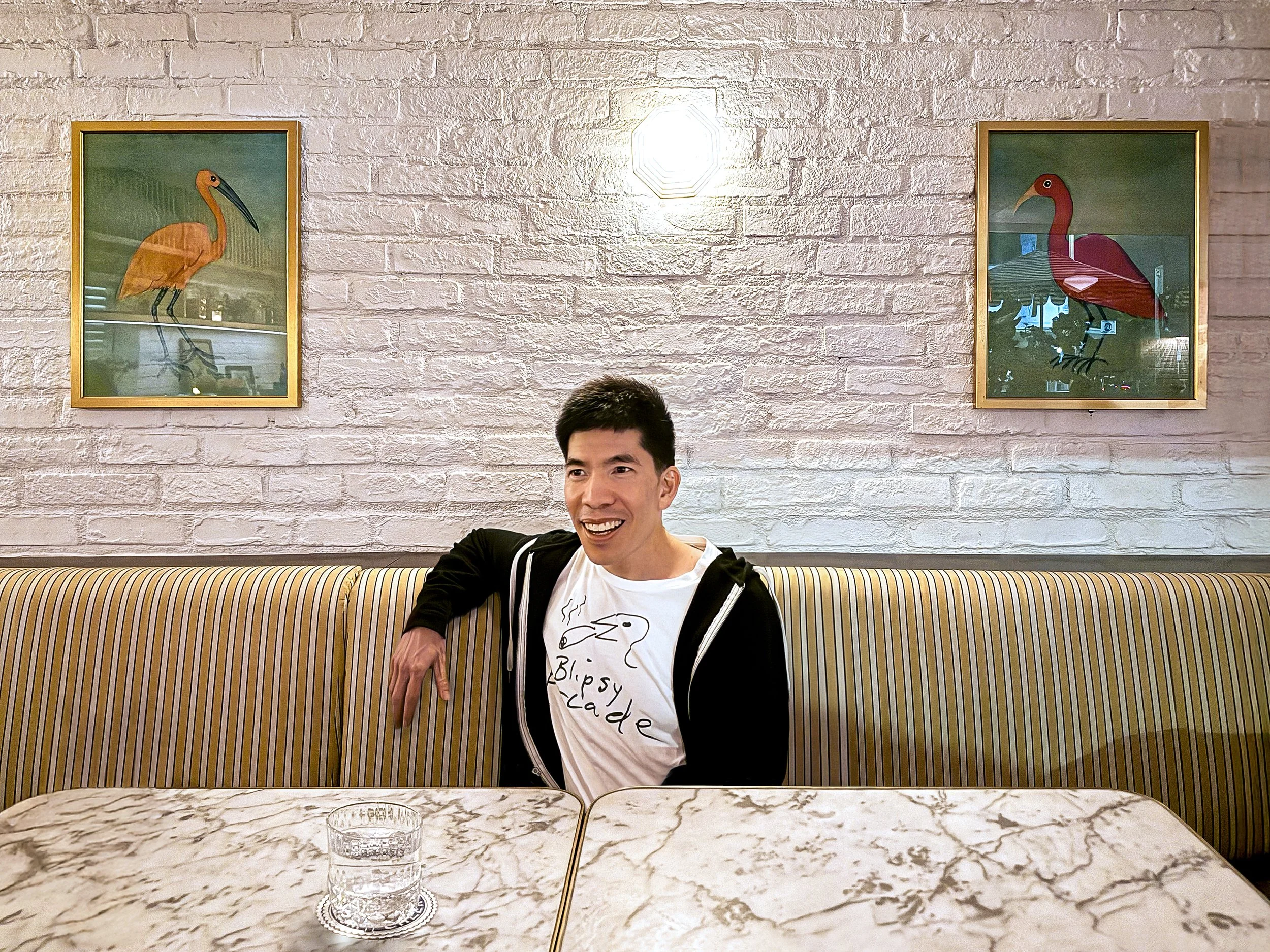 A young man with short dark hair is sitting at a marble table in a restaurant or cafe. He is smiling and wearing a black hoodie over a white T-shirt with a cartoon poodle and the words "Bipsey Cade" printed on it. Behind him, two framed pictures of colorful toucan birds are hanging on a white brick wall. There is a glass of water on the table.