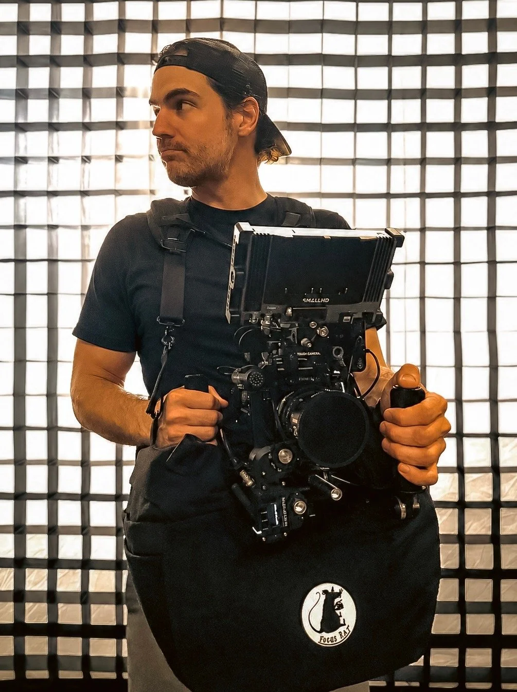 A man with a black baseball cap backwards, black t-shirt, and a harness, holding a professional camera rig, standing in front of a illuminated geometric patterned wall.