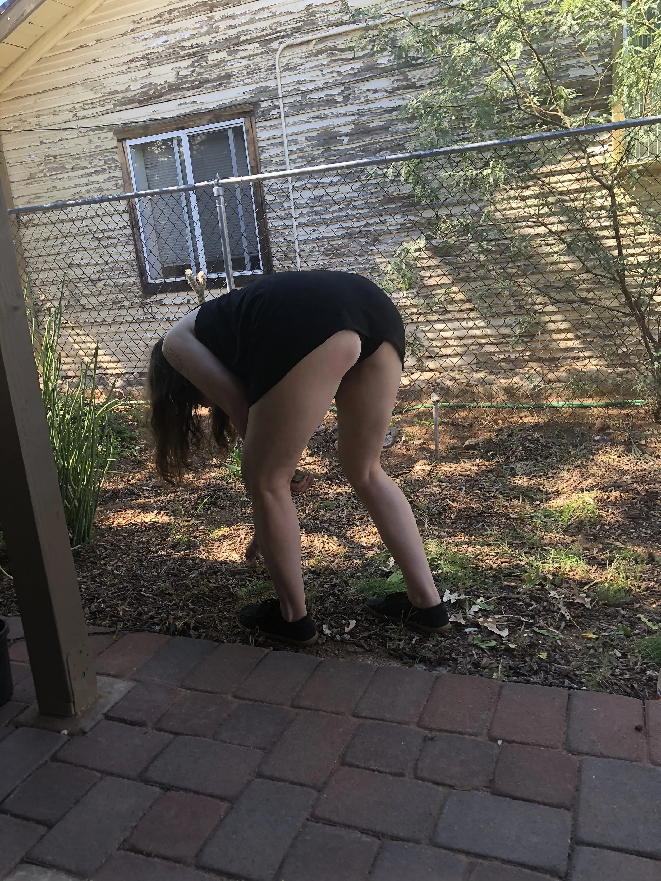 A person with long hair, dressed in black shorts and a black shirt, bending over in a backyard garden area. The background features a wooden house wall with peeling paint, a window, and a chain-link fence with some green foliage.