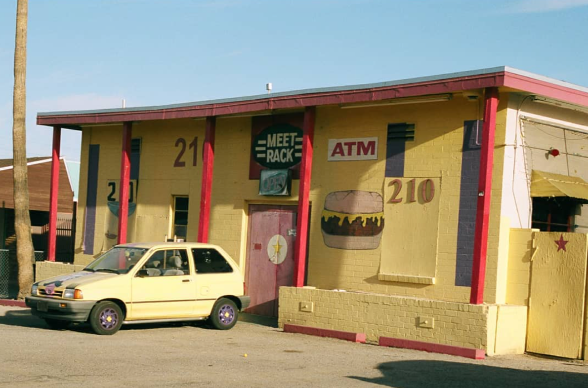 Yellow exterior of Meet Rack with red pillars, featuring painted signs and an ATM sign. There is a painted burger and large numbers on the wall, with a small yellow car parked in front.