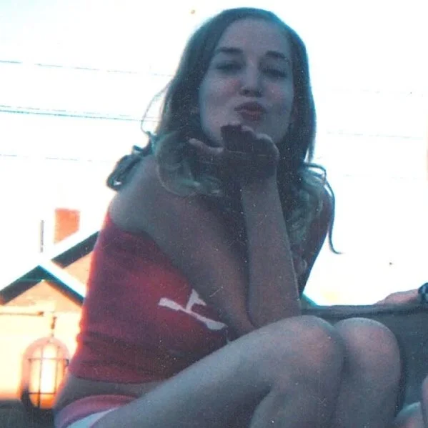 A young woman with dark hair sitting outdoors, blowing a kiss or pucker, wearing a red top.