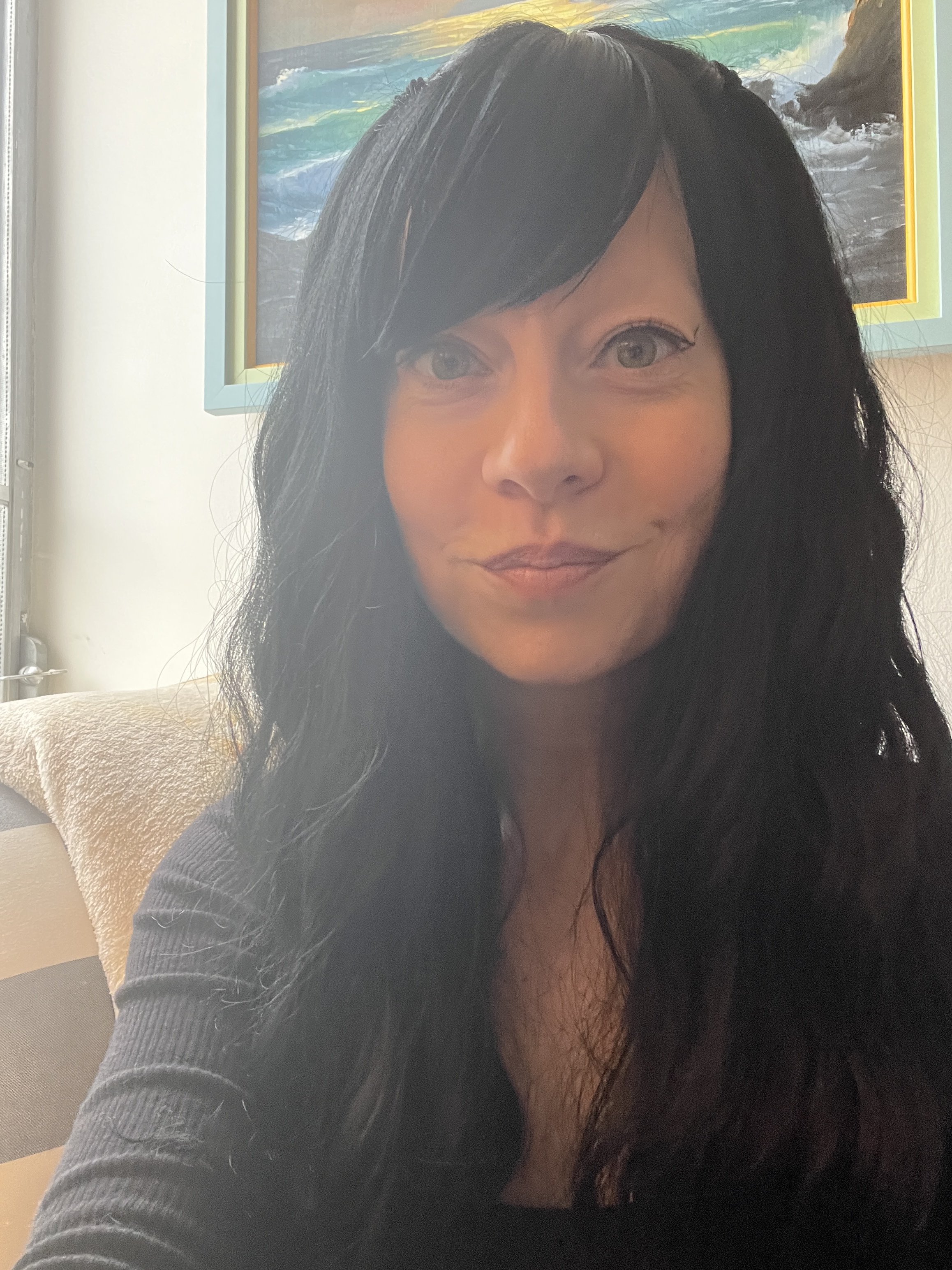 A woman with long dark hair taking a selfie indoors. She is wearing a black top and smiling slightly. There are framed paintings of ocean scenes on the wall behind her.