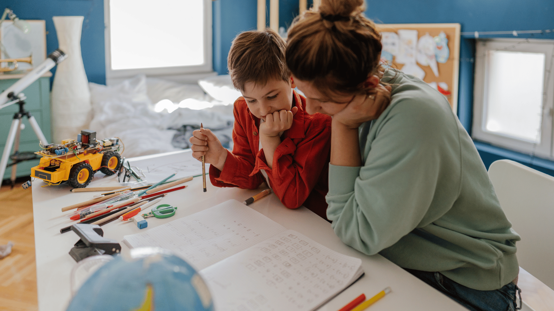 A woman and a boy sit at a white table, looking at a notebook filled with notes and sketches, surrounded by art supplies and a small robotic vehicle, in a brightly lit room with windows and educational posters.