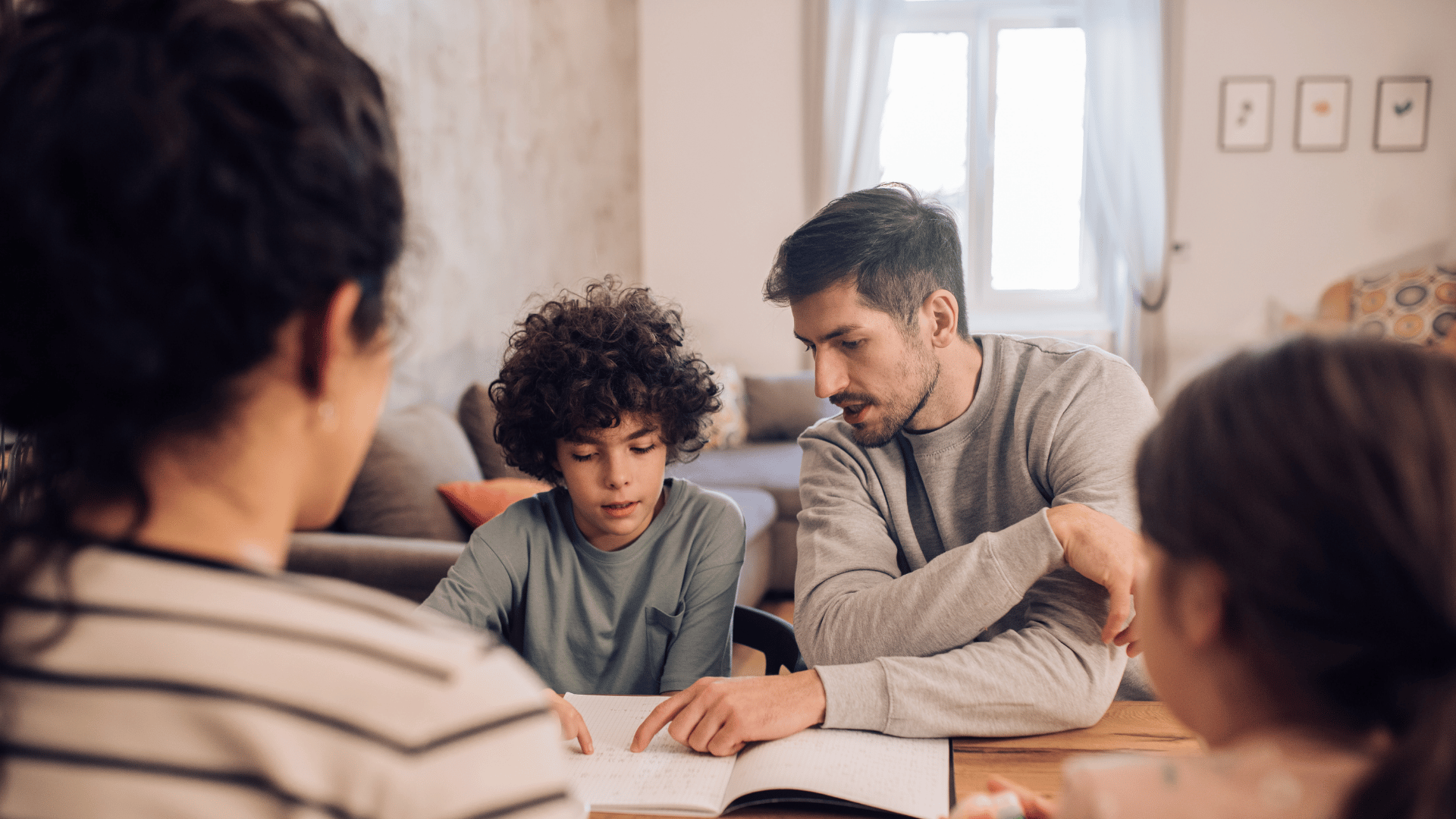 Family sitting around a table in a living room, engaging with a notebook or book, with a woman on the left, a father and son in the middle, and a girl on the right.