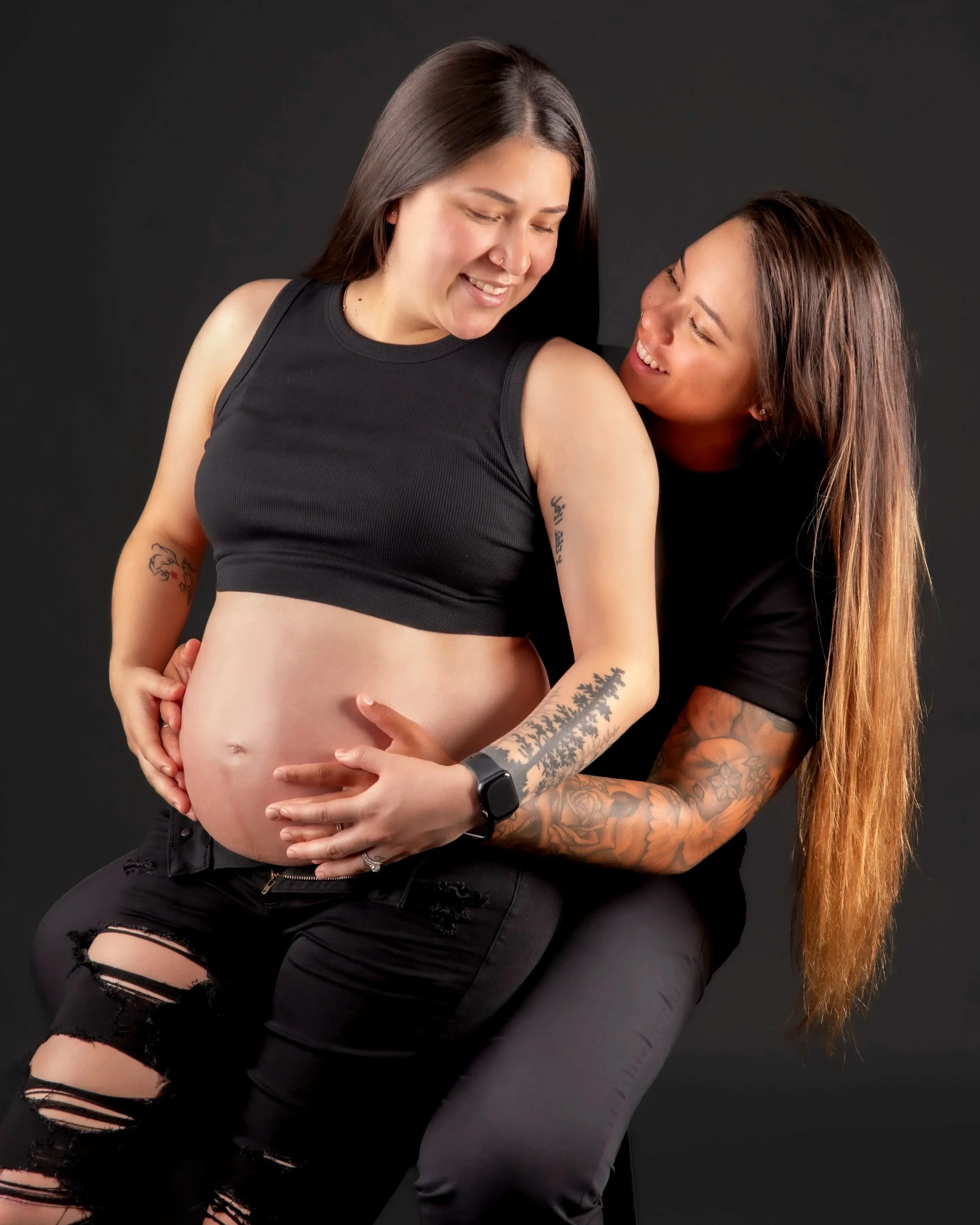 Two women, one pregnant and the other hugging her, smiling and looking at each other. They are wearing black clothing with ripped details, against a dark background.