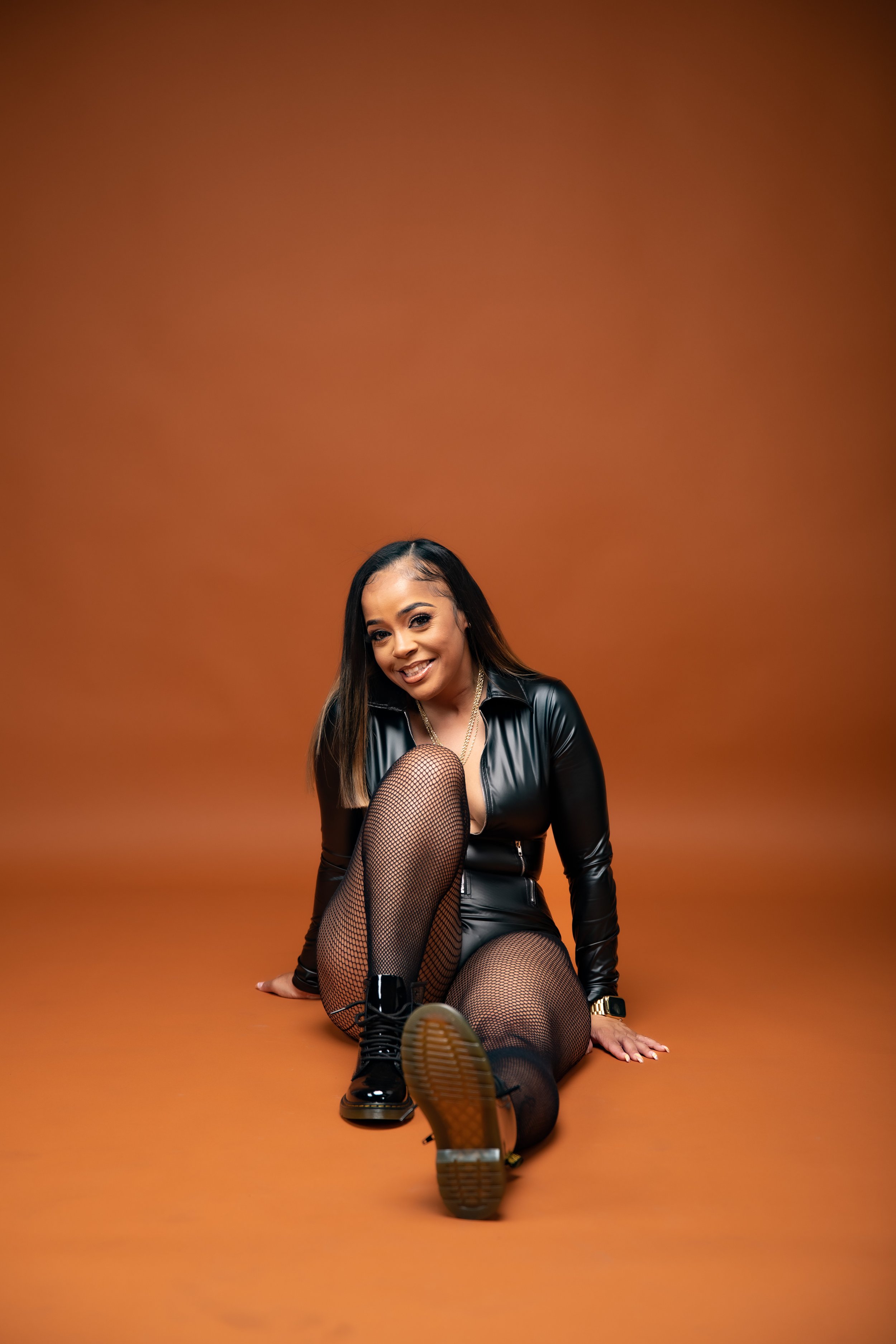 A woman with long dark hair smiling, wearing a black leather jacket, fishnet tights, and combat boots, sitting on the floor against an orange backdrop.