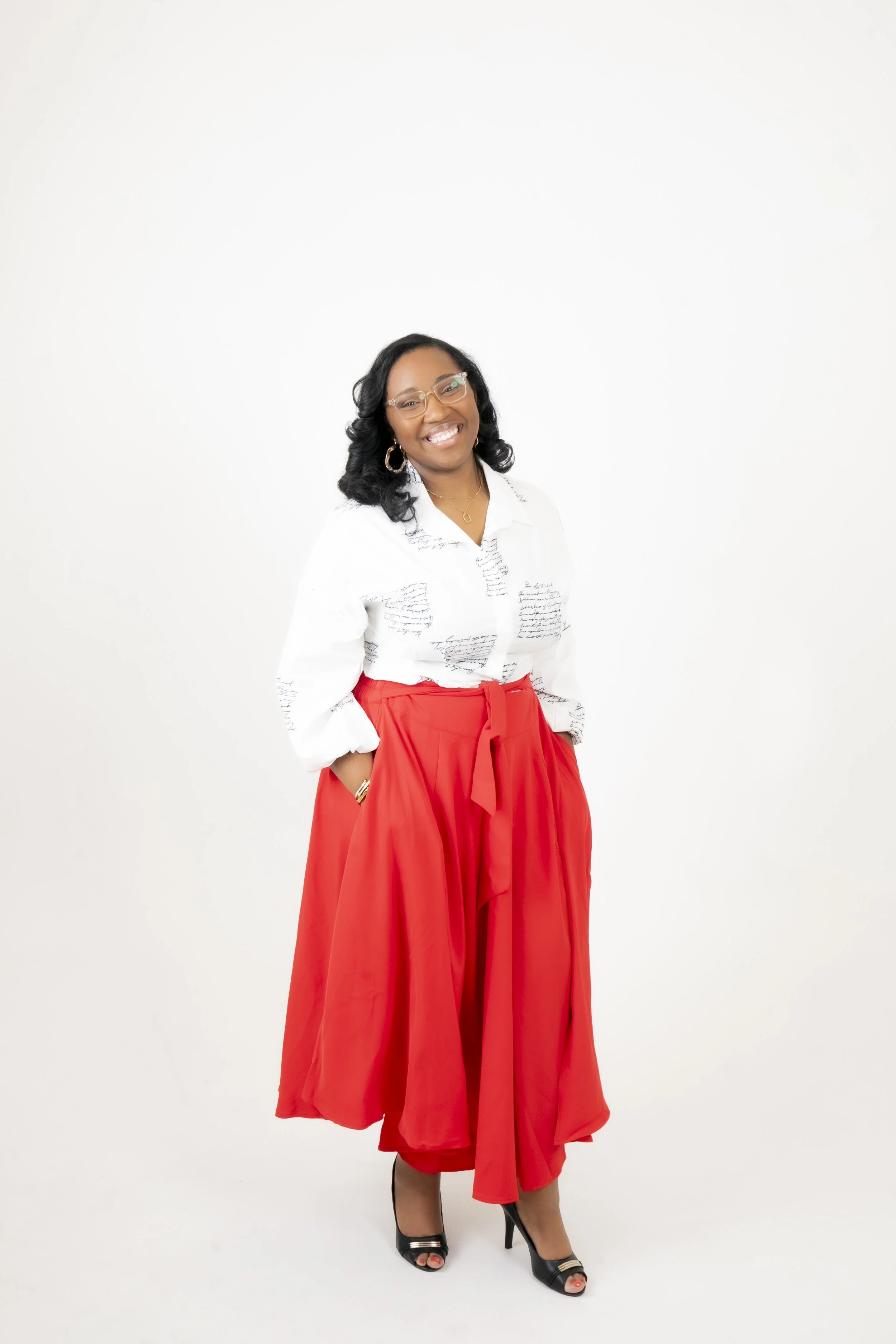 A woman smiling, wearing glasses, a white shirt with black script text, red wide-leg pants tied at the waist, and black high-heeled shoes, standing against a plain white background.