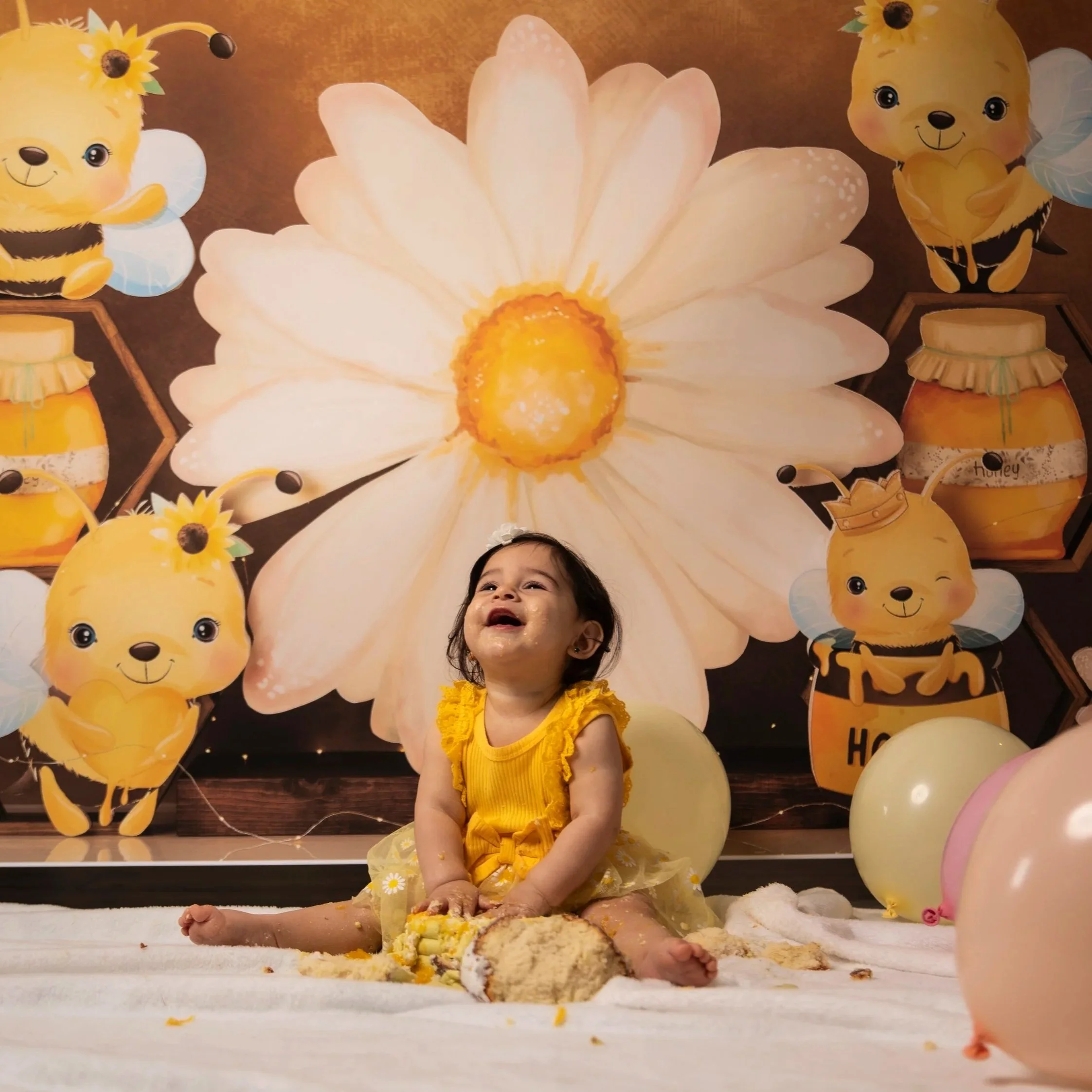 A young girl in a yellow dress with daisies, sitting on a table covered in cake and frosting, surrounded by helium balloons with bumblebee and flower designs, and large flower and bee decorations on the wall.
