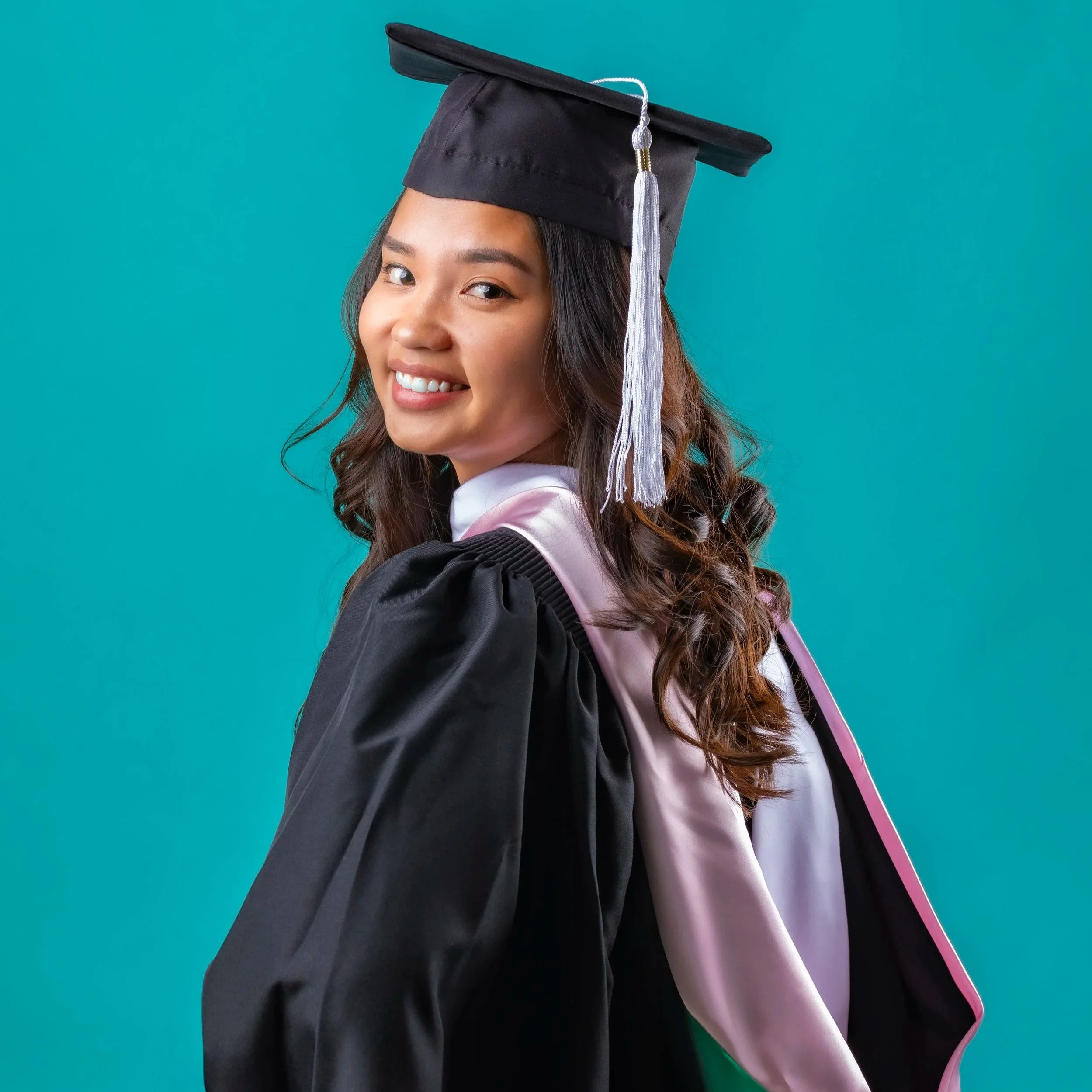 A young woman wearing a black graduation cap and gown smiling at the camera with a turquoise background.