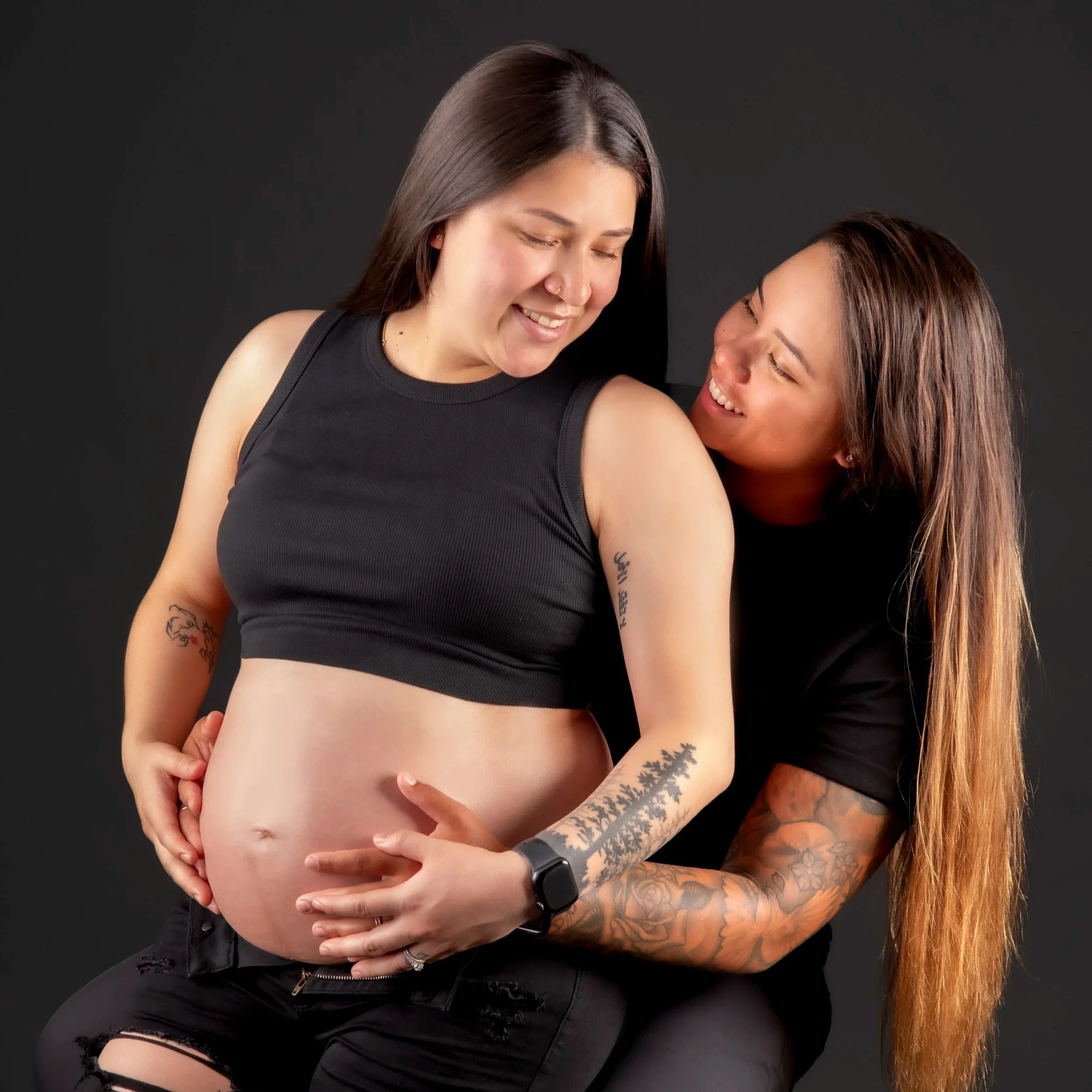 Two women, smiling and hugging against a black background. One has straight, dark brown hair and a nose piercing, wearing a black sleeveless top. The other has long, wavy brown hair and tattoos on her arm, wearing a black T-shirt. The woman with straight hair is holding the waist of the woman with tattoos.