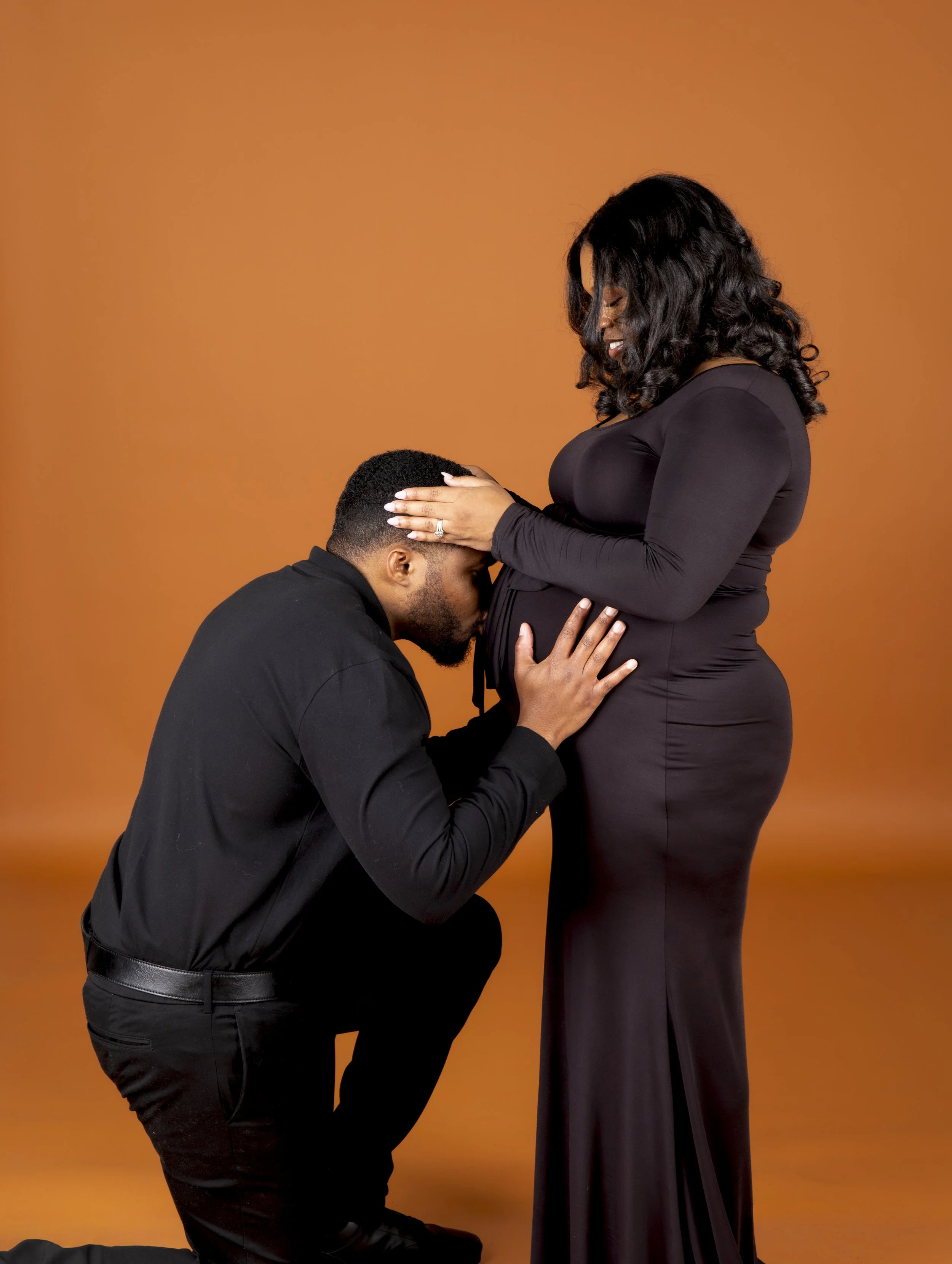 A man kneeling and holding a pregnant woman's belly, while the woman gently rests her hands on her pregnant belly and smiles. Both are dressed in black, standing against an orange background.