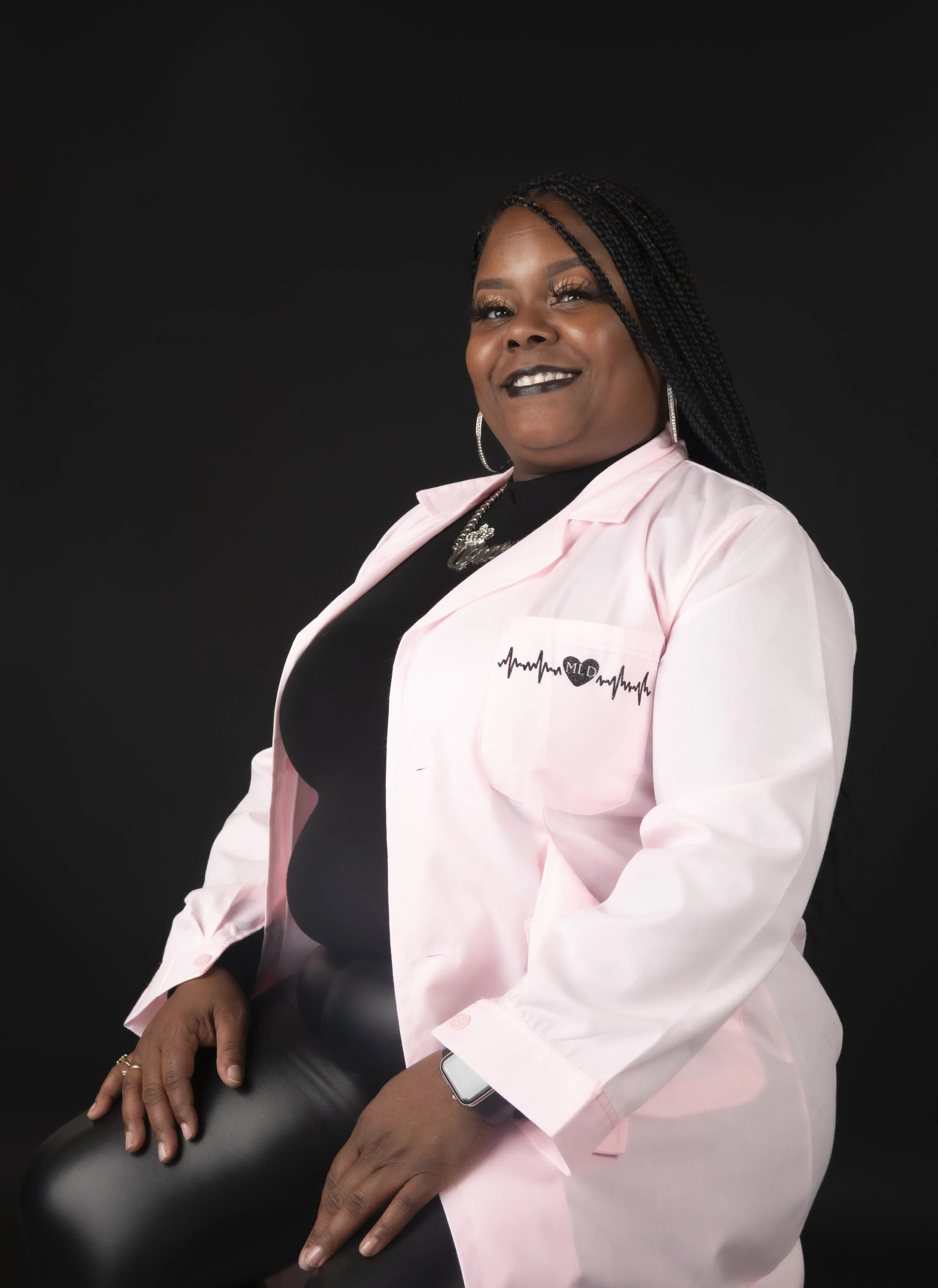 Portrait of a smiling woman with braided hair wearing a pink jacket with a medical logo, black top, and jewelry against a black background.